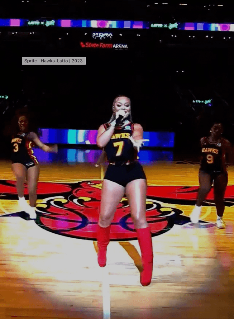 Latto wearing a Hawks jersey and red boots sings on a basketball court, with two backup dancers behind her, during a halftime show at State Farm Arena.