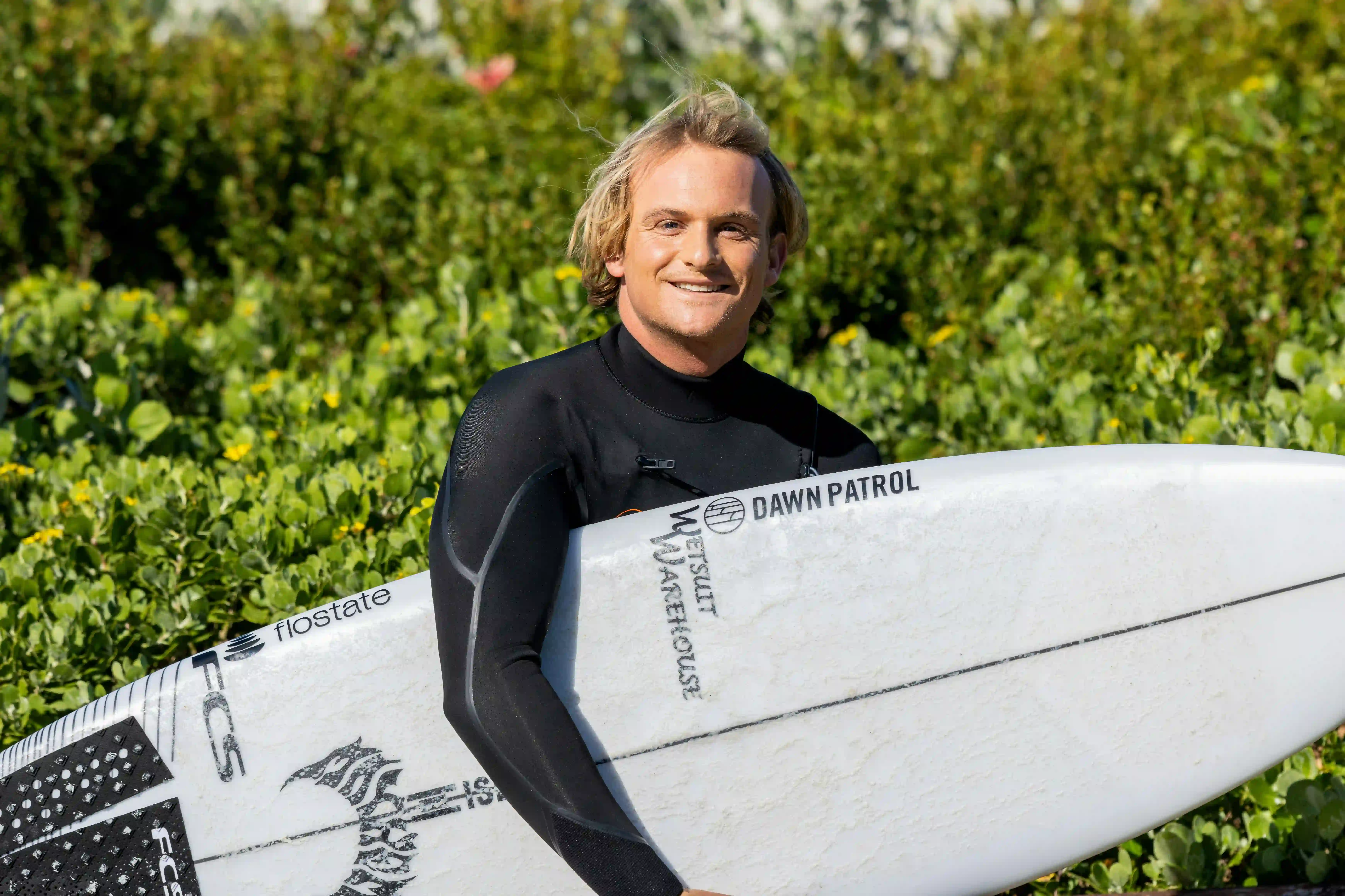 Smiling surfer in a black wetsuit holding a white surfboard in front of green bushes.