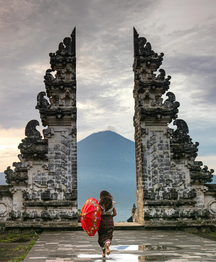 A person with a red umbrella walks through a temple gate toward a mountain.