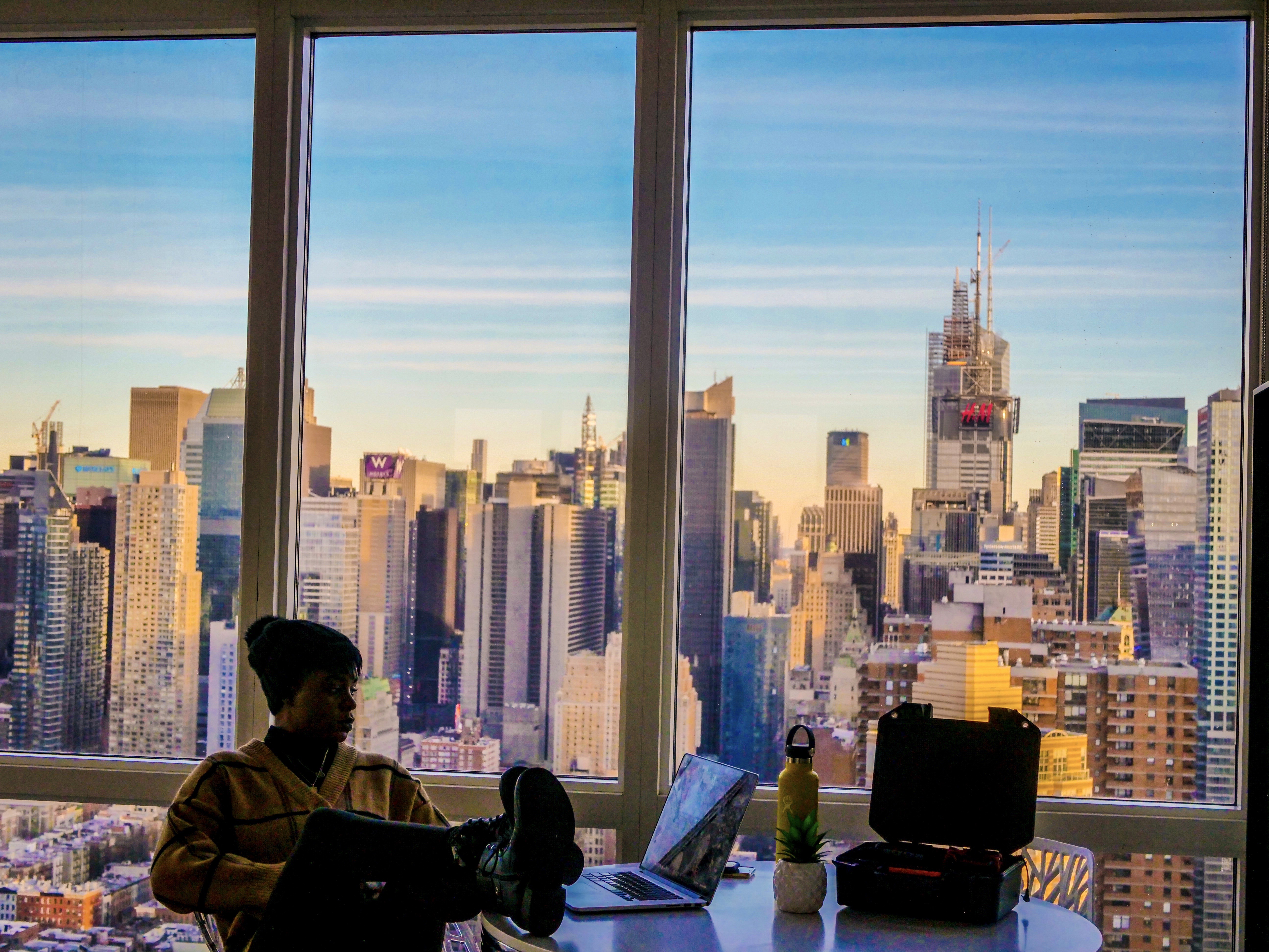 A person sits near a window with a city skyline at sunset, featuring tall buildings and a colorful sky.