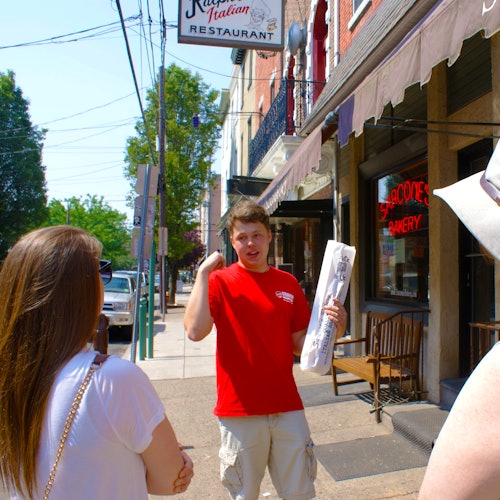 A person in a red shirt speaks to two people on a city sidewalk near an Italian restaurant and bakery.