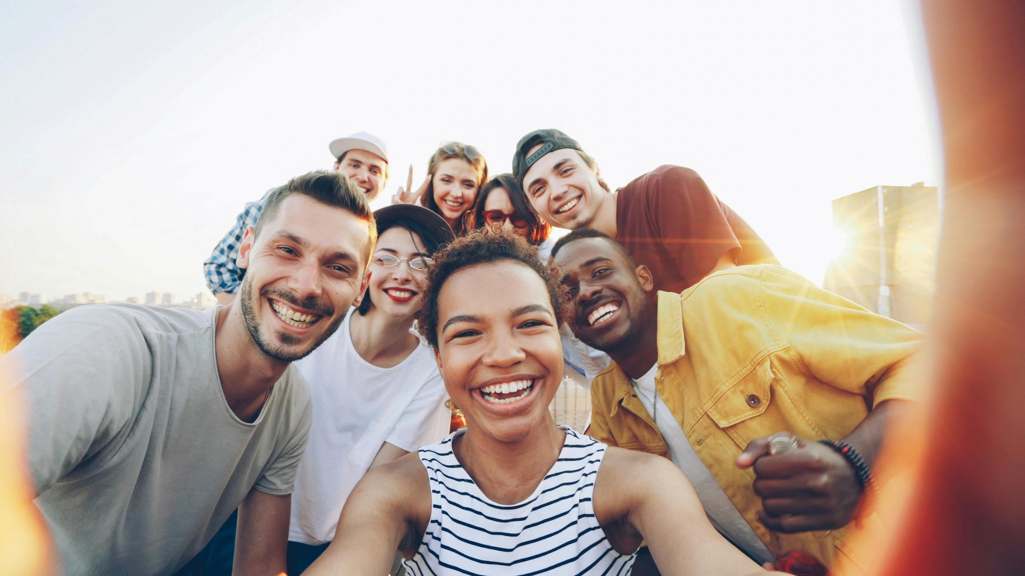 A vibrant, wide-angle selfie of a diverse group of seven young adults smiling together outdoors on a sunny day. In the center foreground, a Black woman with curly hair holds the camera, laughing in a striped tank top. Grouped tightly around her are several friends—including a White man in a grey t-shirt, a White woman in a black hat, and a Black man in a yellow shirt—all grinning warmly. The bright, overexposed sky and warm lens flare create a cheerful, candid, and energetic atmosphere.
