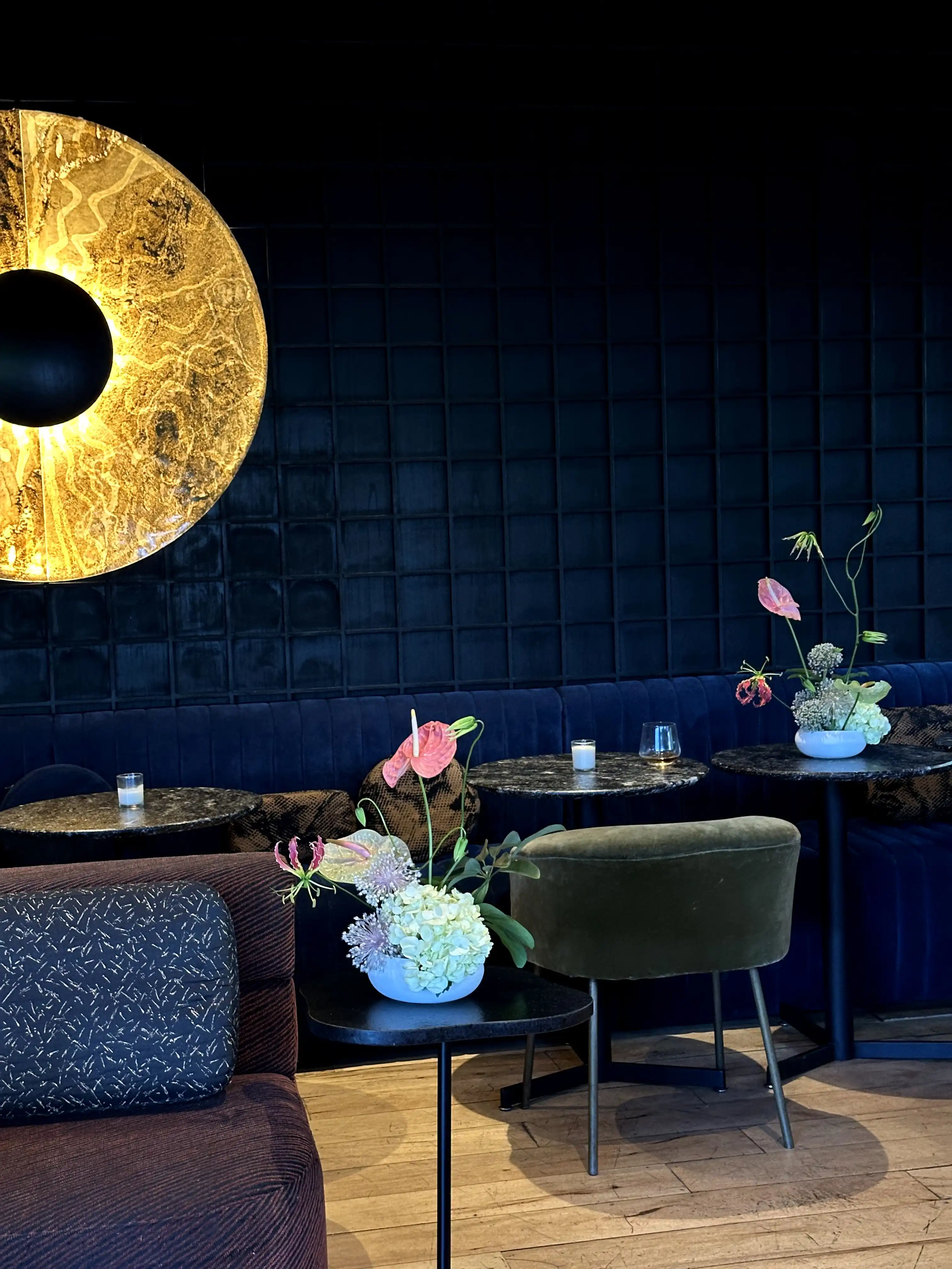 Interior elegante con paredes oscuras y lámpara dorada circular iluminada. En las mesas de mármol negro destacan arreglos florales con anturios rosados, hortensias y gloriosas, acompañados de velas pequeñas y sillones aterciopelados en tonos verdes y azules.