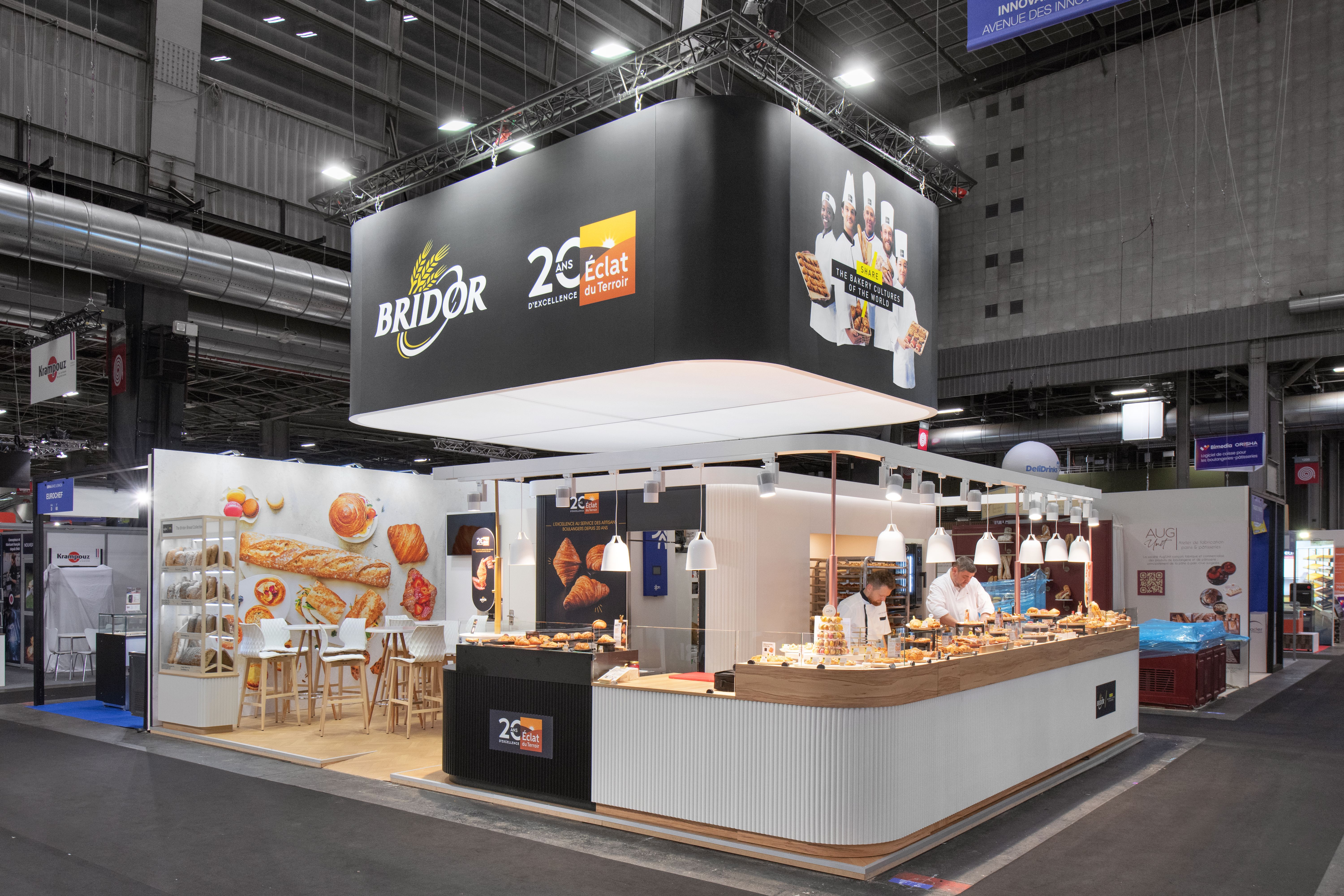 Stand Bridor Bake and Snack 2026 réalisé par Expace – vue frontale avec caisson anthracite suspendu logo et 20 ans Éclat du Terroir, comptoir dégustation boulangerie et espace lounge produits