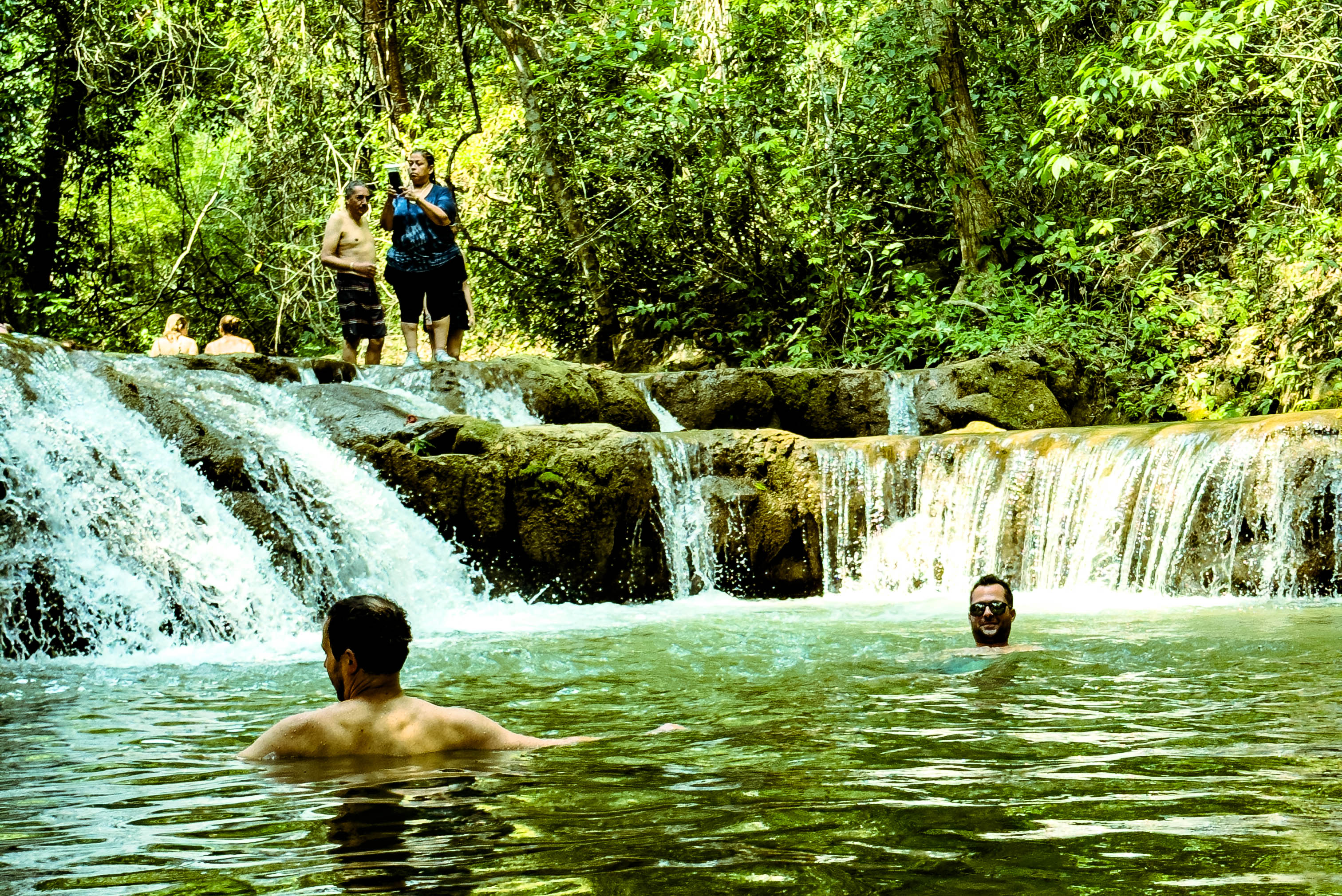 people swimming and standing in a waterfall in the middle of the jungle