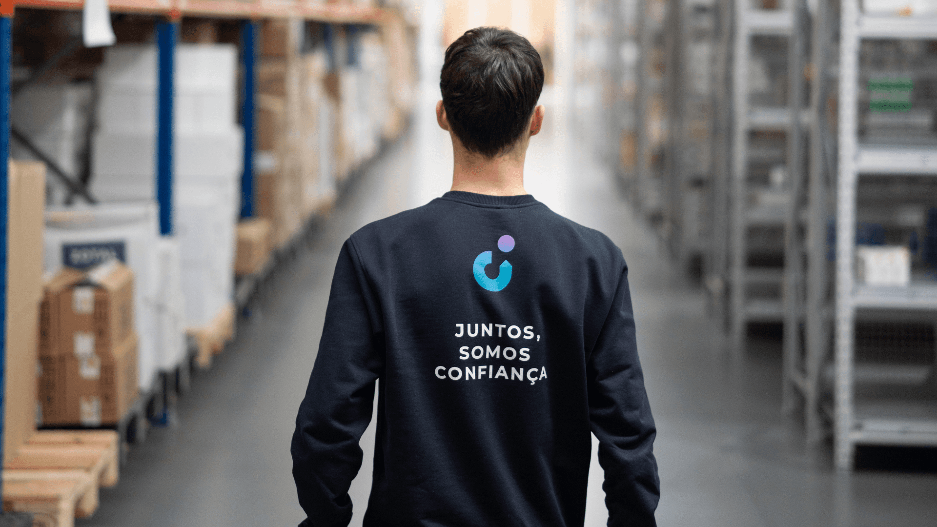 Pessoa de costas em um armazém de distribuição, vestindo uma camiseta com a frase 'Juntos, somos confiança', promovendo segurança e colaboração logística.
