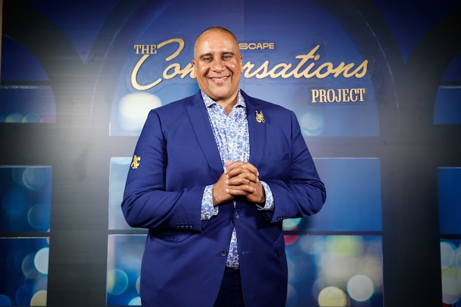 A smiling Marc Spears in a blue suit stands in front of a backdrop that reads The Conversations Project, with blurred blue and yellow lights in the background.