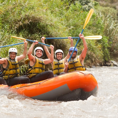 Four people in helmets and life vests celebrate while white-water rafting in an orange inflatable boat on a river.