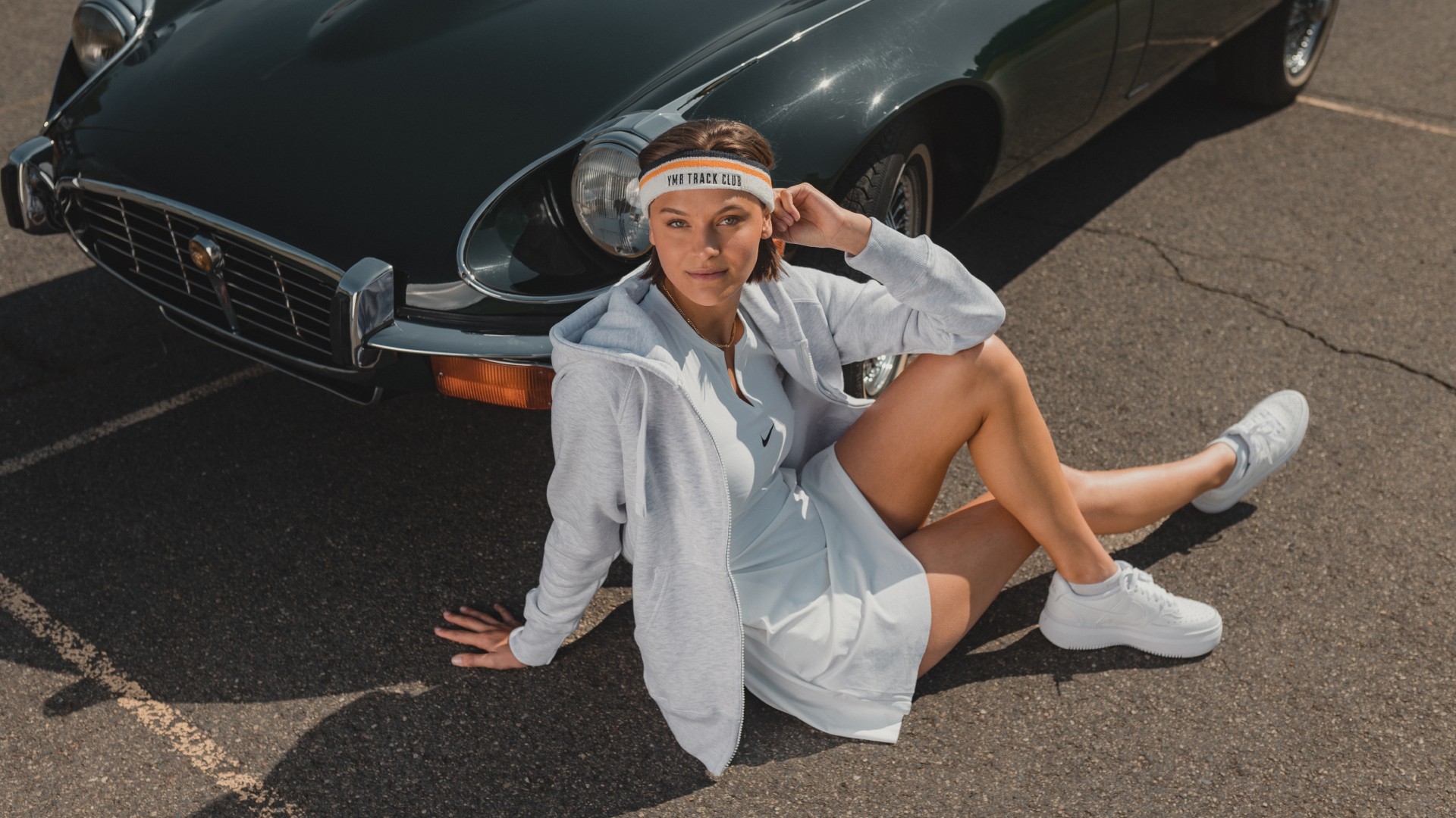 A young woman dressed in a white dress and light grey ziphoodie, sitting on the ground in front of a green sportscar.
