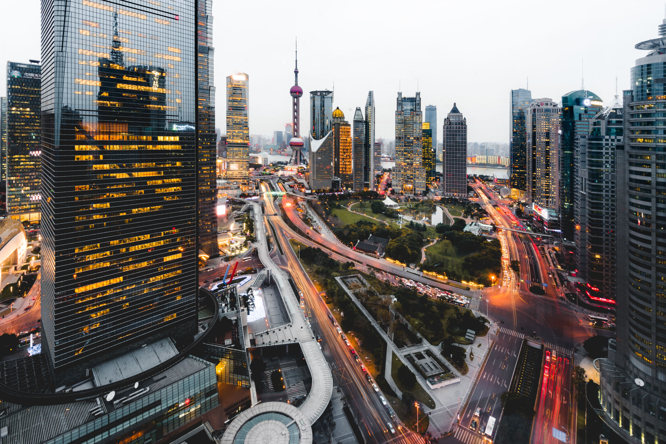 High-angle evening view of a busy city intersection with skyscrapers, glowing lights, and traffic trails near a tower.