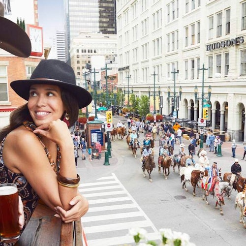 A woman in a black hat smiles while watching a horse parade from a balcony in a city with tall buildings.