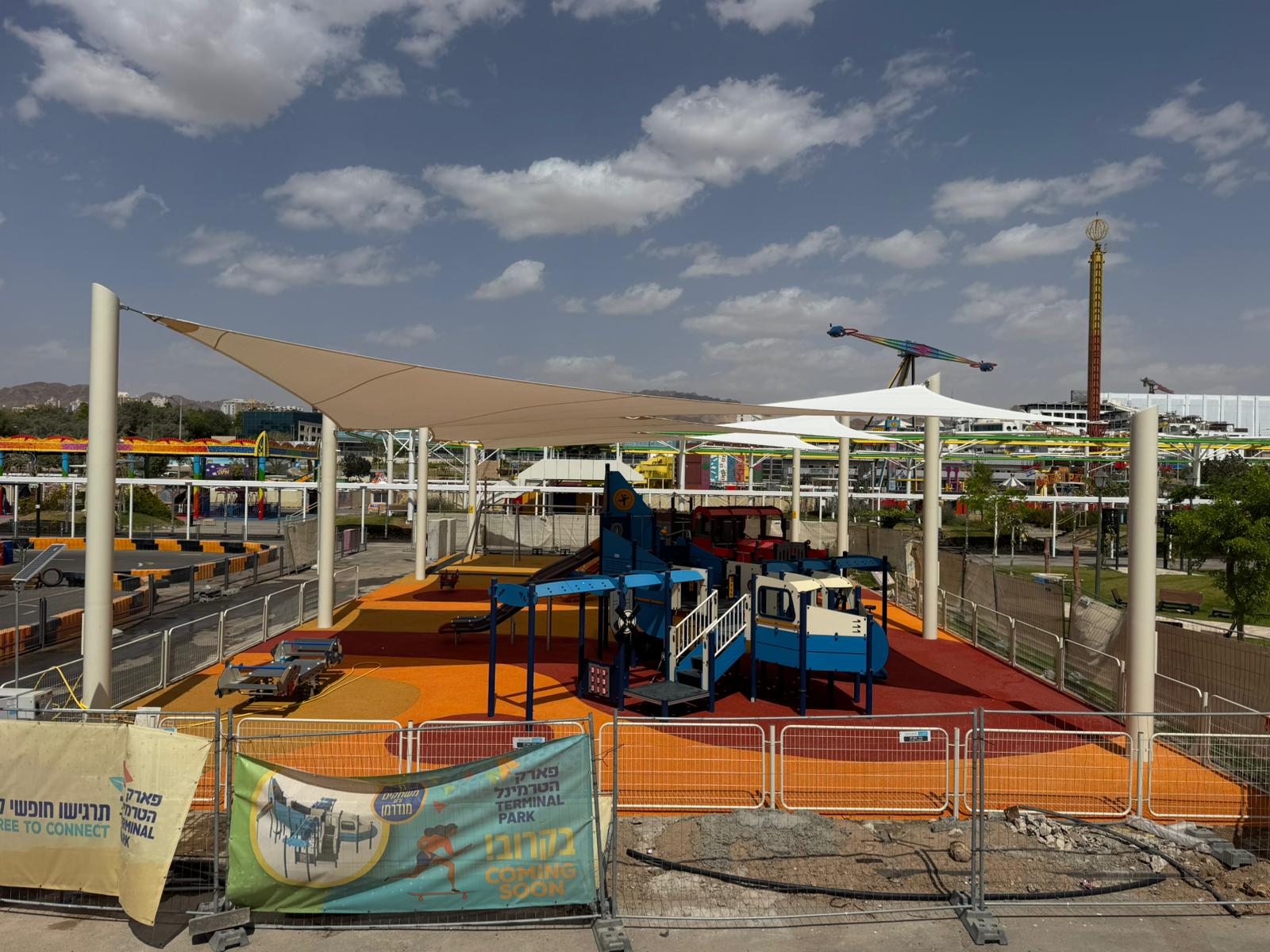 Playground with climbing structure and shade sails surrounded by construction fences with amusement park rides in the background.