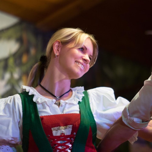 Uma pessoa em um traje tradicional com uma blusa branca, colete verde e espartilho vermelho, sorrindo enquanto segura as mãos com outra pessoa.
