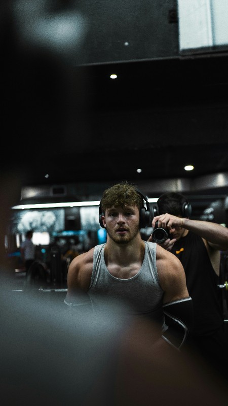 A man in a white tank top standing at a squat rack. Quote: “The strength training plan helped me recover from a sports injury…” – Former College Athlete.
