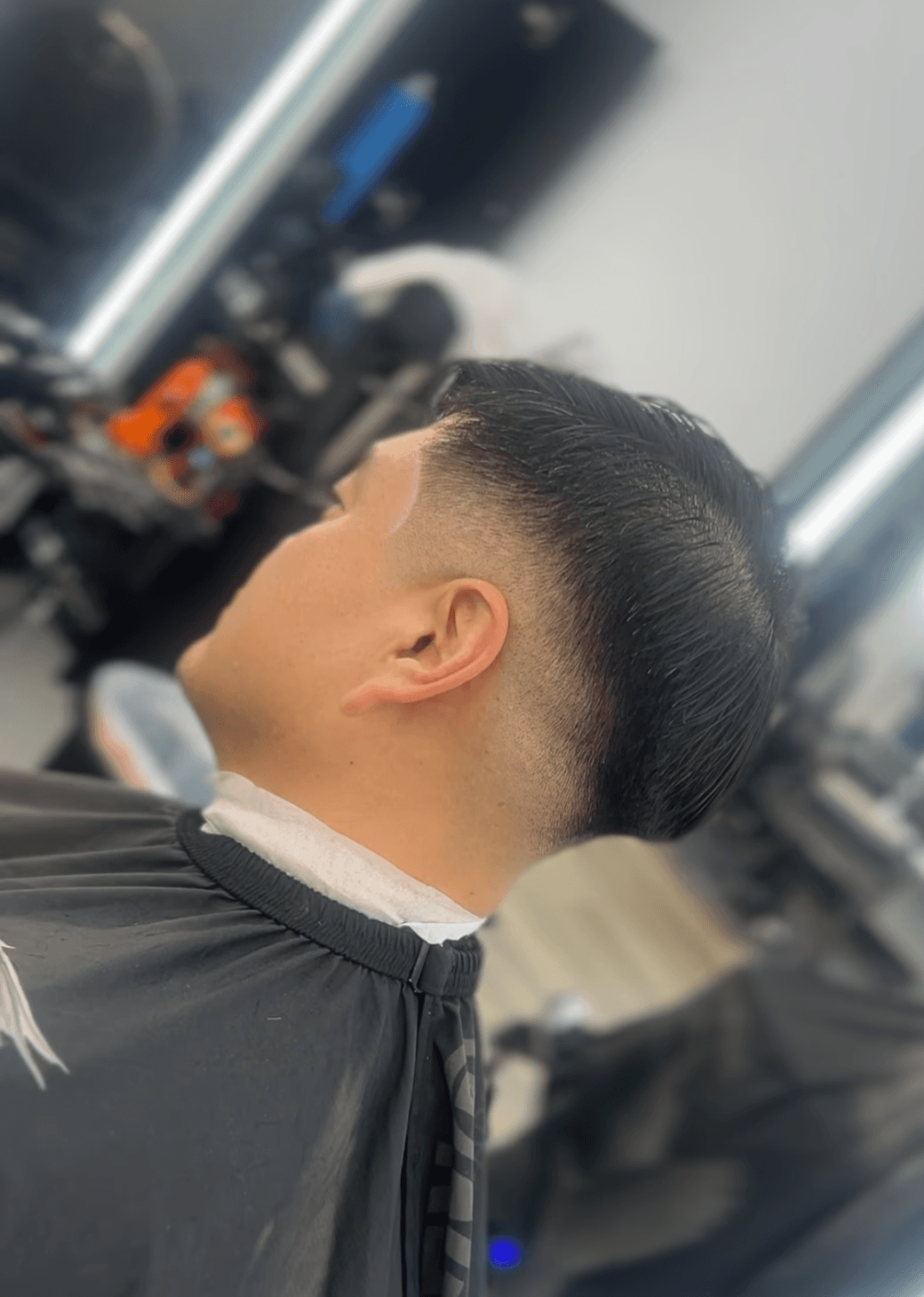 Man sitting in a barber chair with a clean fade haircut, short top, and straight fringe while looking at his phone.
