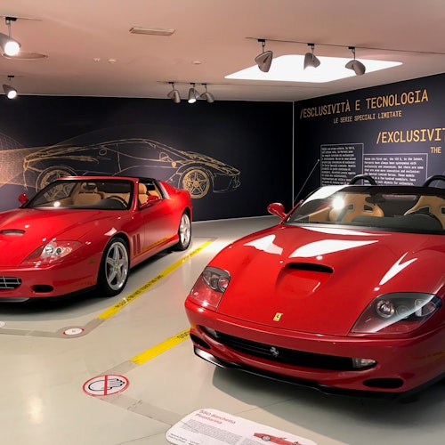 שני מכוניות פרארי פתוחות אדומות מוצגות באולם תצוגה עם לוחות מידע ותאורה מלמעלה.
