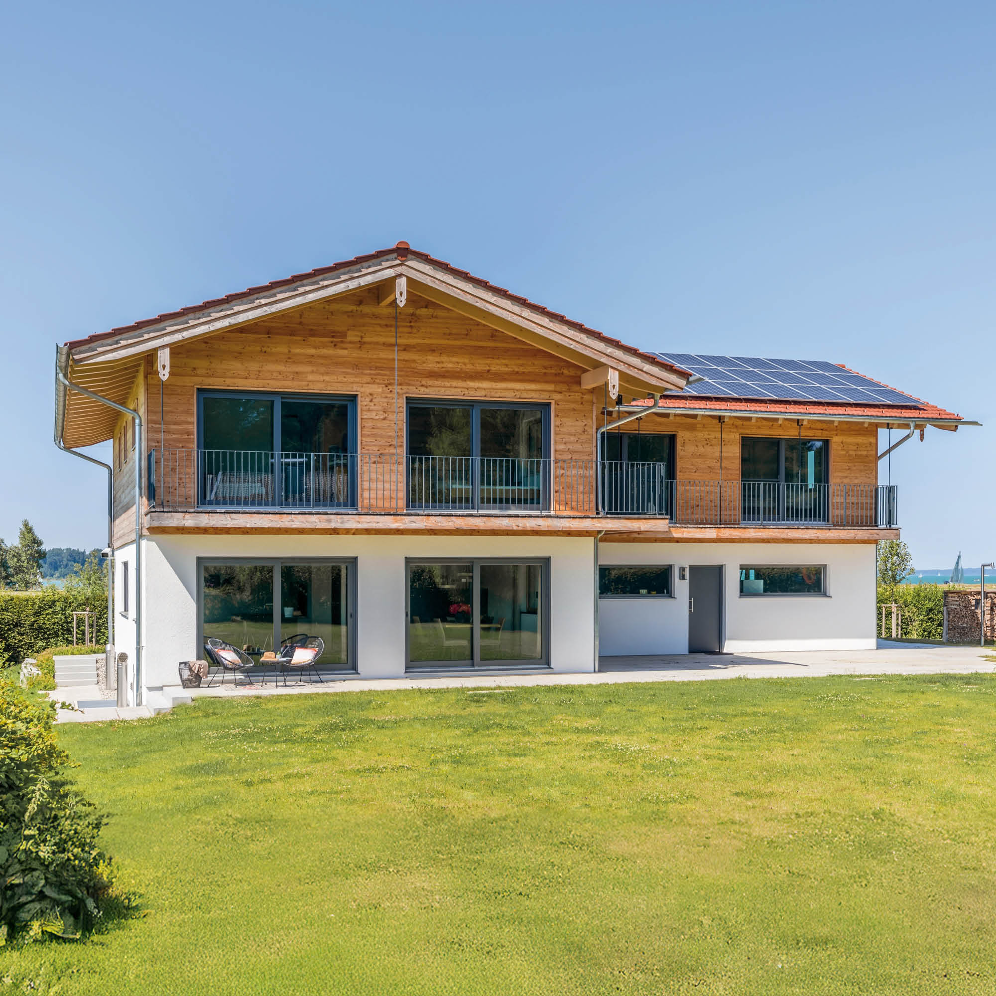 Modernes Holzhaus mit Solaranlage auf dem Dach, großen Fensterfronten und Balkonen, gelegen in einer grünen Landschaft bei Tageslicht.