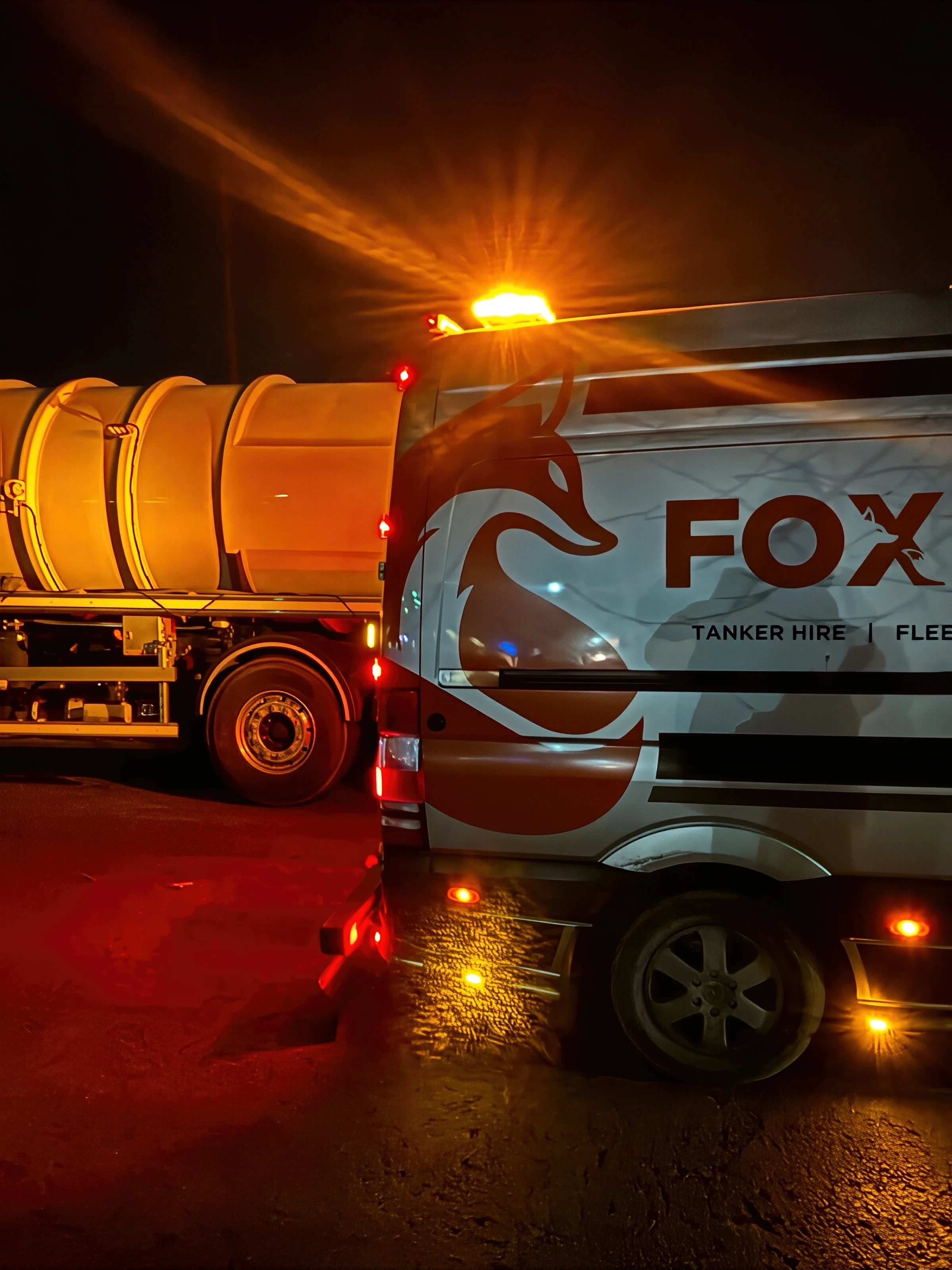 Nighttime close-up of a Fox-branded truck with amber hazard lights on, parked near a tanker trailer under an orange streetlight.