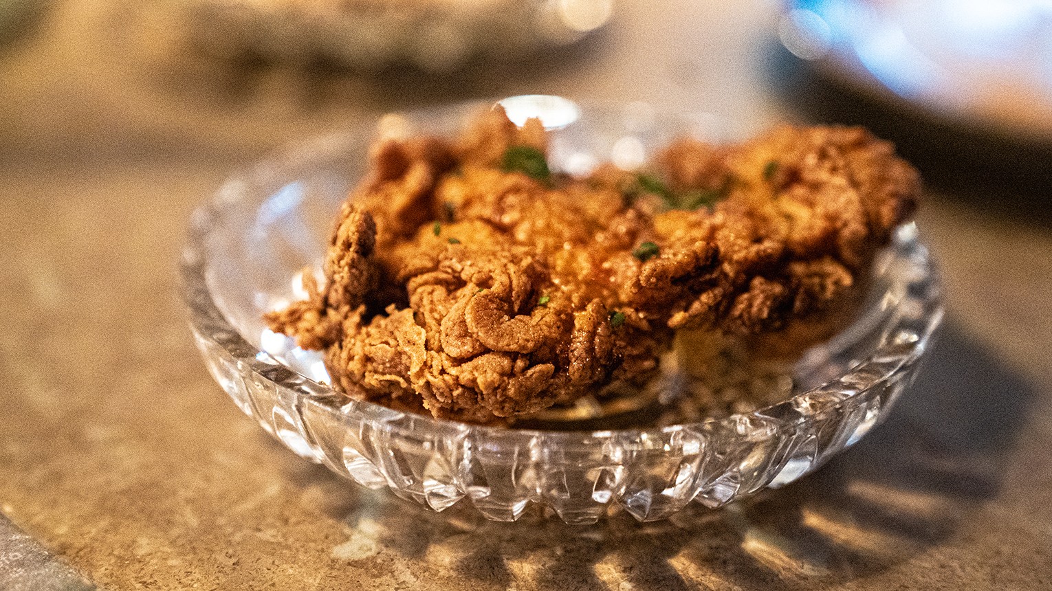 A woman in sunglasses and jeans crouches with a suitcase on a sunny, rural path