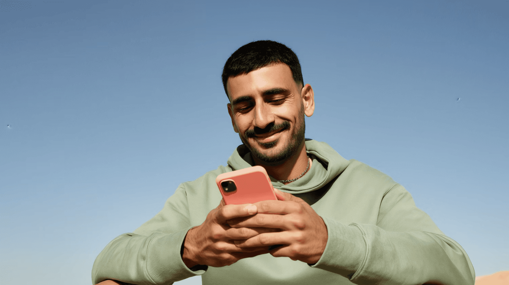A man in a green hoodie smiles while using a coral-colored smartphone under a clear blue sky.