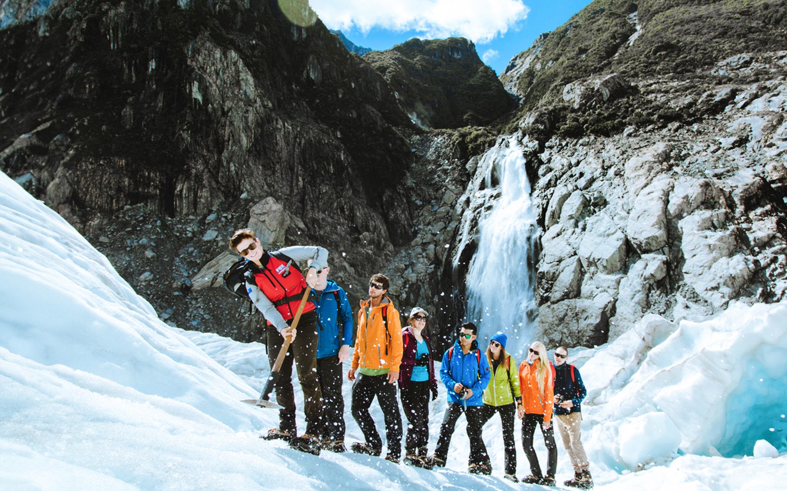 Grupo de excursionismo en el glaciar Fox, Nueva Zelanda, con una cascada al fondo.