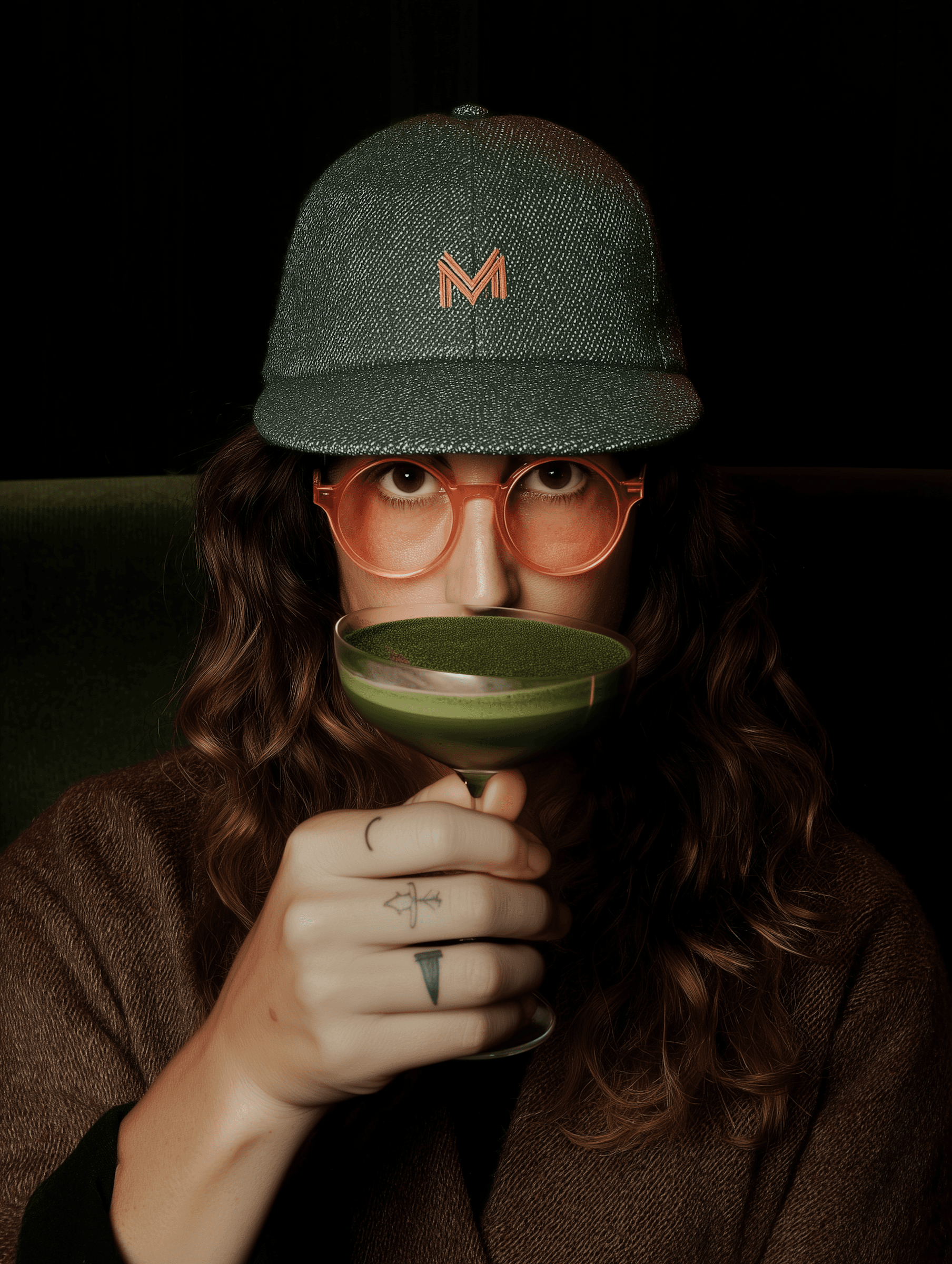 A stylish individual with a green cap and orange glasses holds a coupe glass of green matcha, showcasing unique tattoos and a cozy vibe.