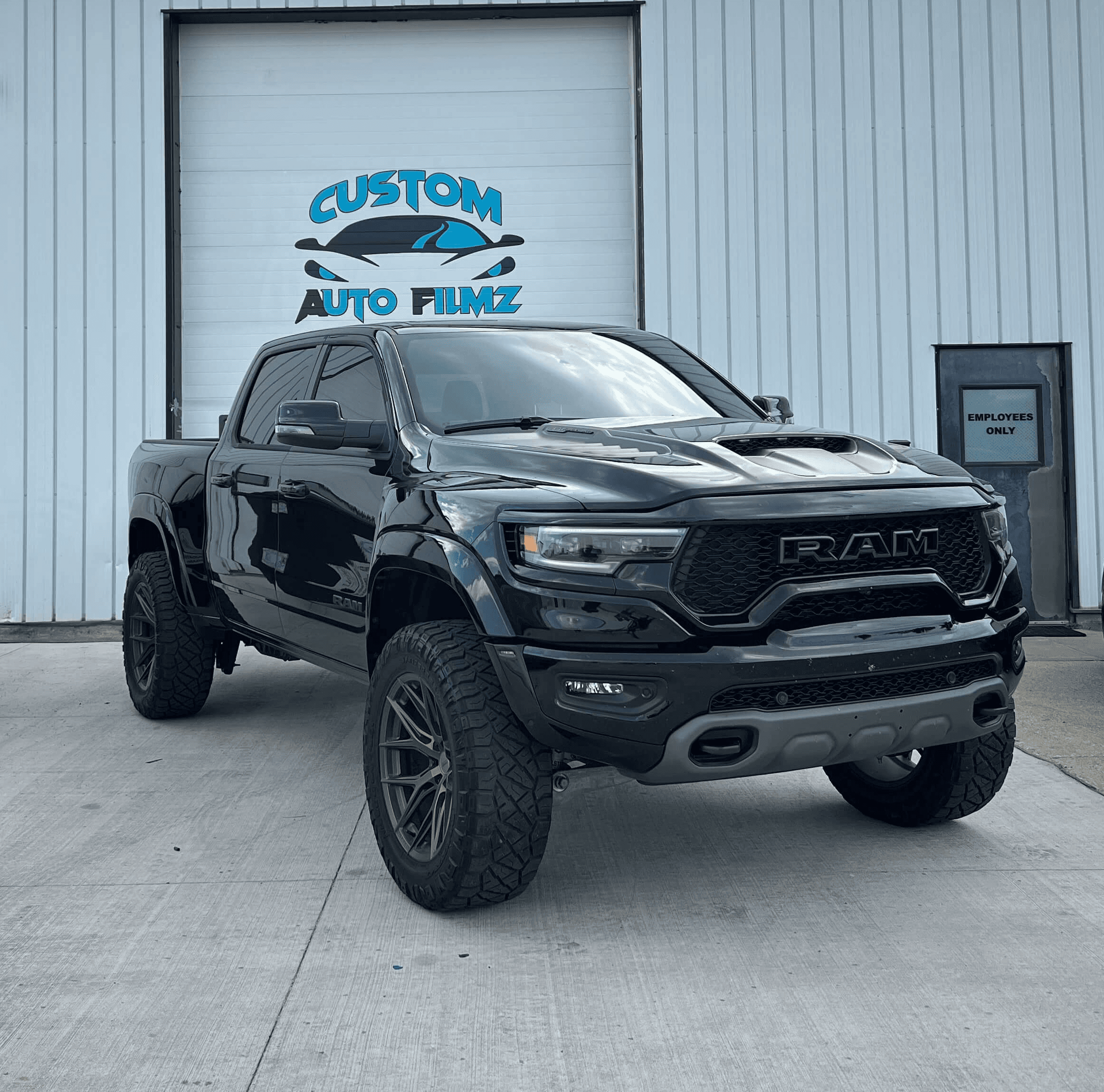 A black RAM 1500 TRX pickup truck is parked on a concrete driveway in front of a white industrial building.