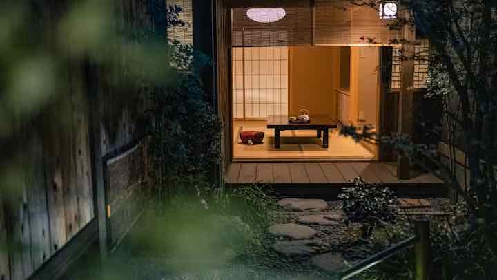 A serene Japanese tea room with wooden interiors, featuring a low table set with a traditional teapot, viewed through a lush garden path under ambient lighting.