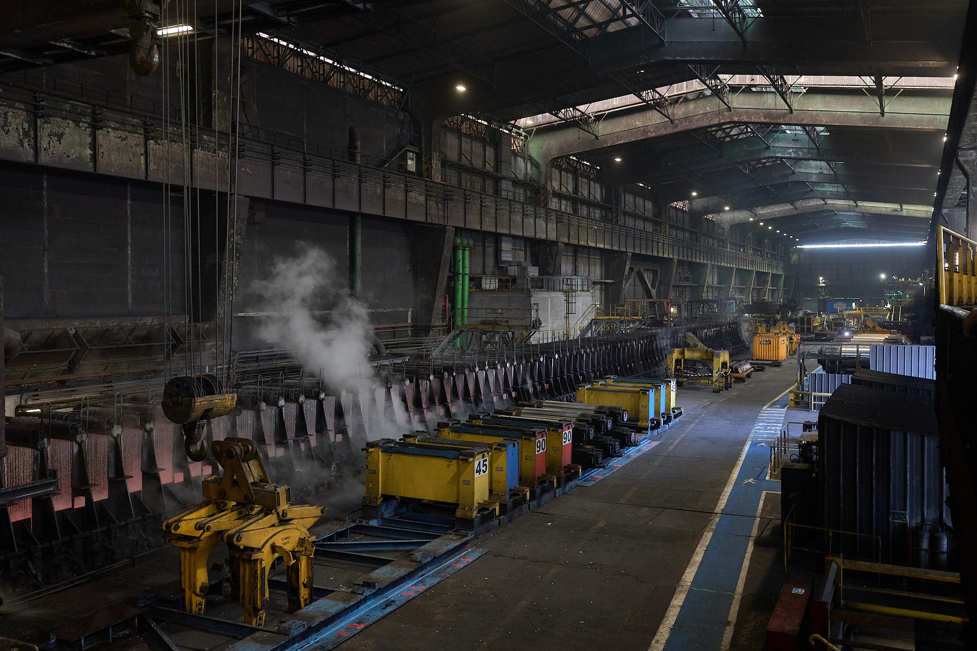 Vue grand-angle d'un hangar industriel ArcelorMittal, avec de la vapeur d'eau qui émane des brames d'acier refroidi par des jets d'eau.