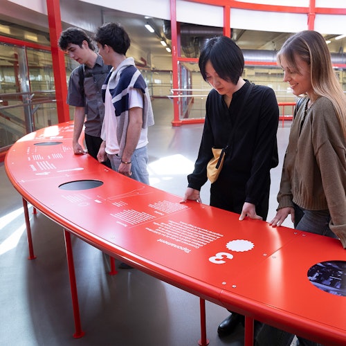 四个人站在一座现代化建筑内，一张弯曲的红色信息展示台旁。建筑内有红色的结构元素。