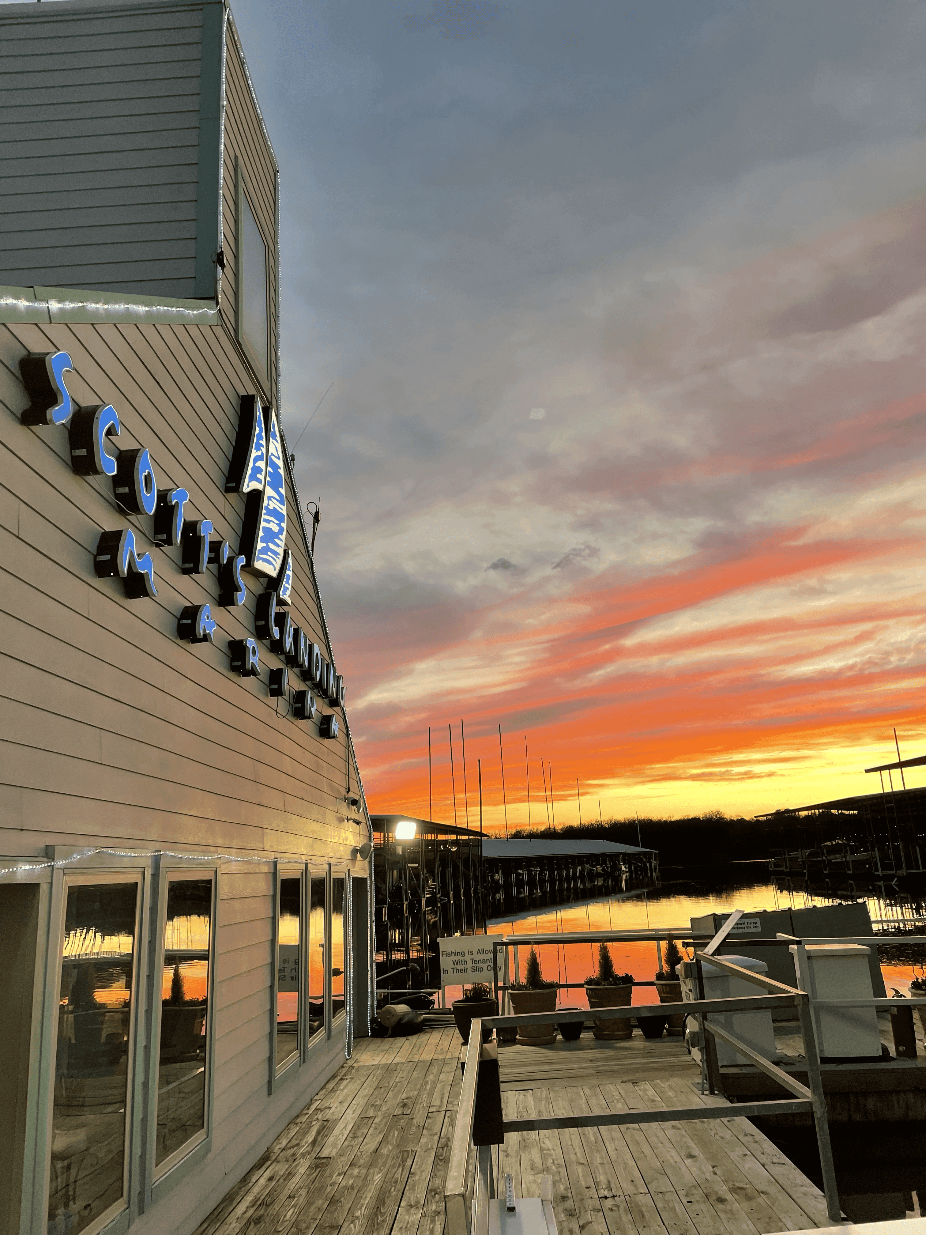 A stunning sunset over a tranquil marina, with vibrant orange and pink hues reflecting in the calm water next to a building bearing a prominent neon sign and rustic wooden dock.