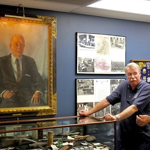 A man stands next to a glass display case with historical artifacts, in front of a large portrait and framed historical photographs on the wall.