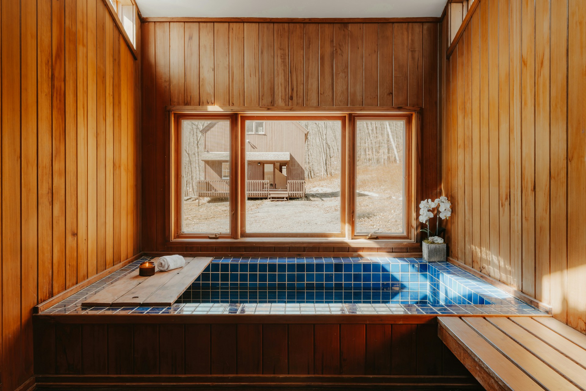 Sunlit wood-paneled room with a blue-tiled tub, window view of a cabin, and orchids. Candle and towel on a wooden board.