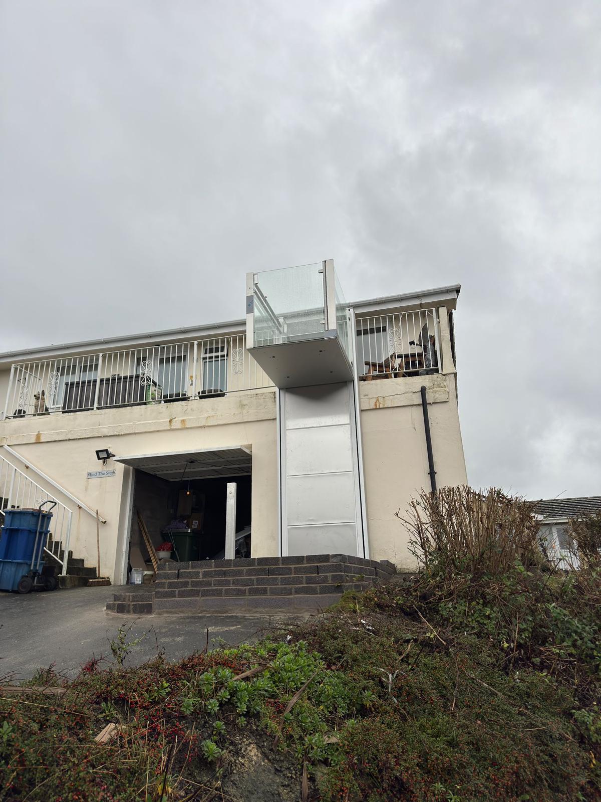 Vertical platform step lift installed on the exterior of a two-storey coastal residential property — full installation view showing column, raised platform at balcony level, and entrance steps below