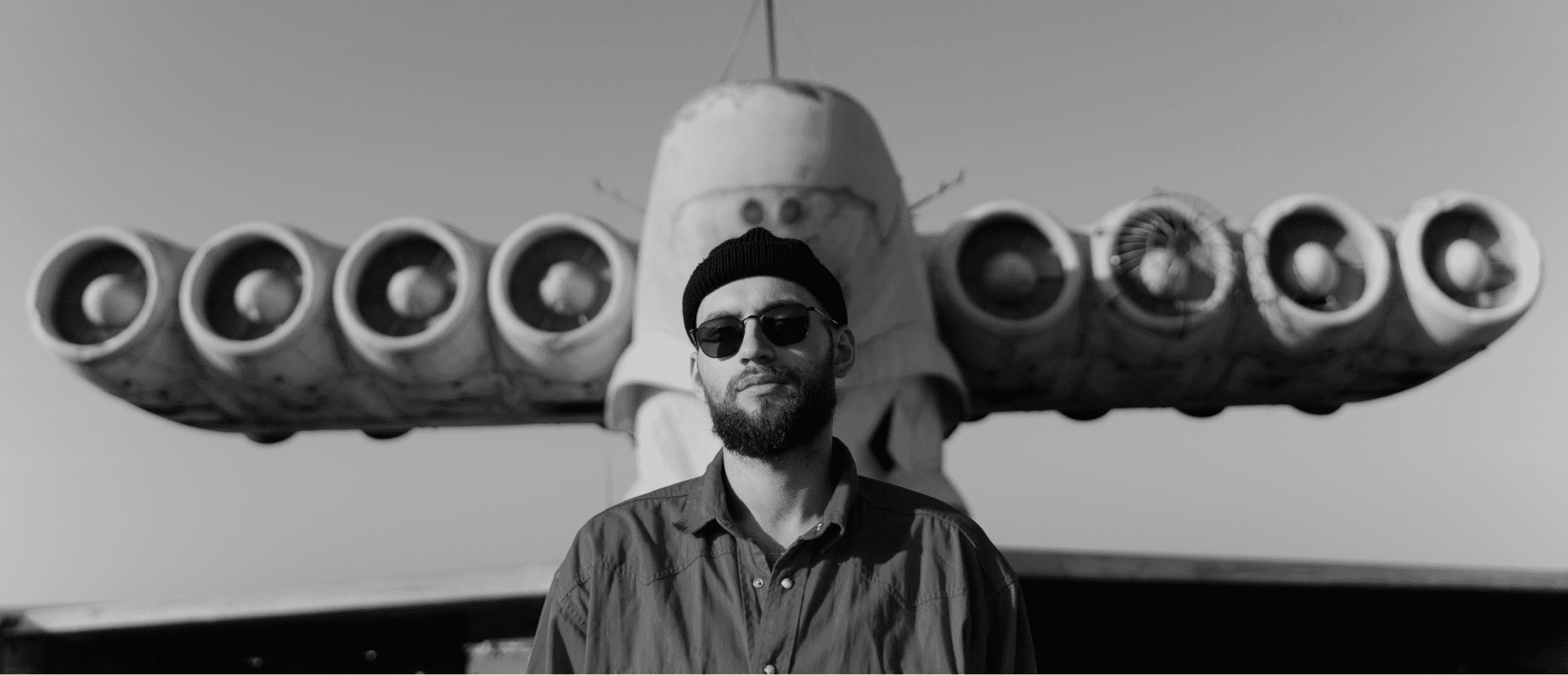 A bearded man wearing a knit cap and sunglasses stands facing the camera, positioned directly in front of the engine housing of an old aircraft.
