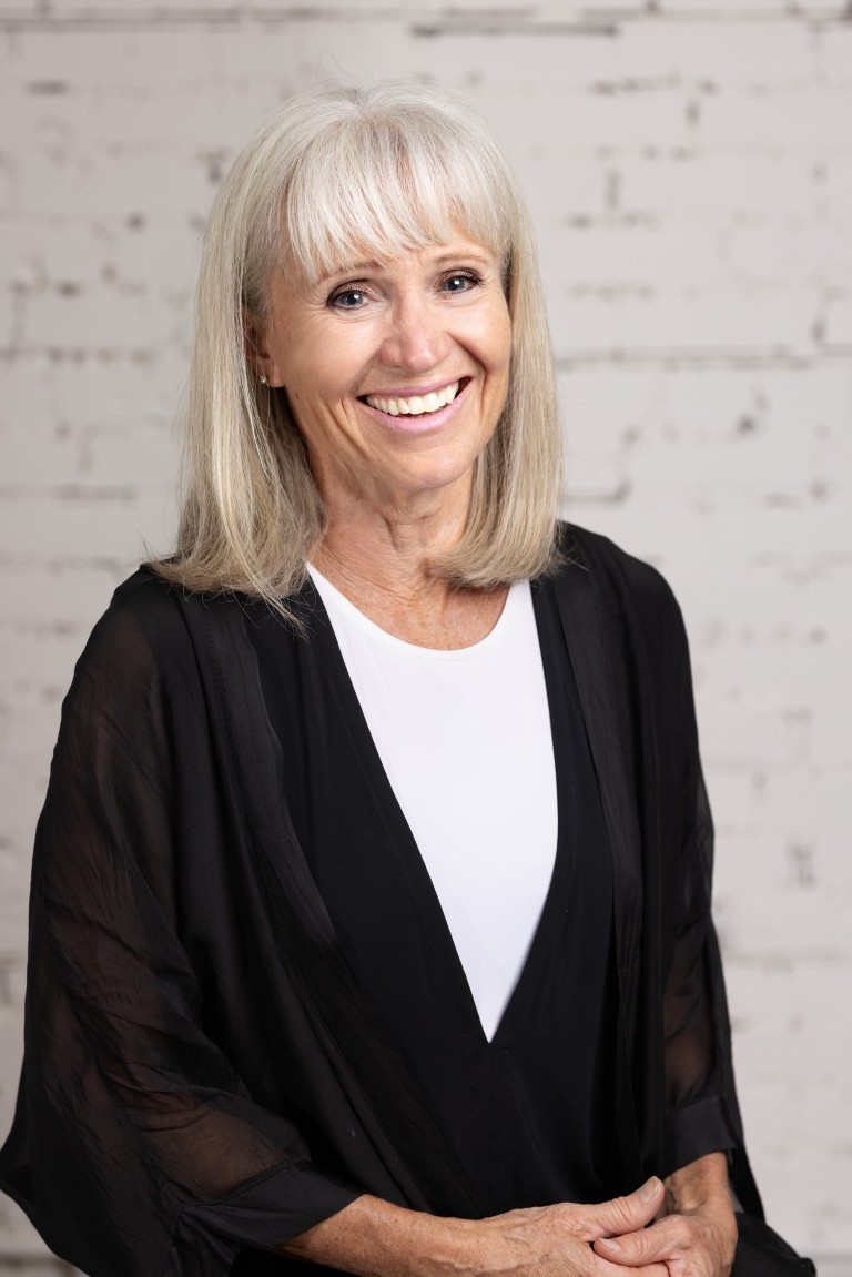 Image of a fair skinned woman with silver hair smiling at the camera with her hands clasped in front of her.