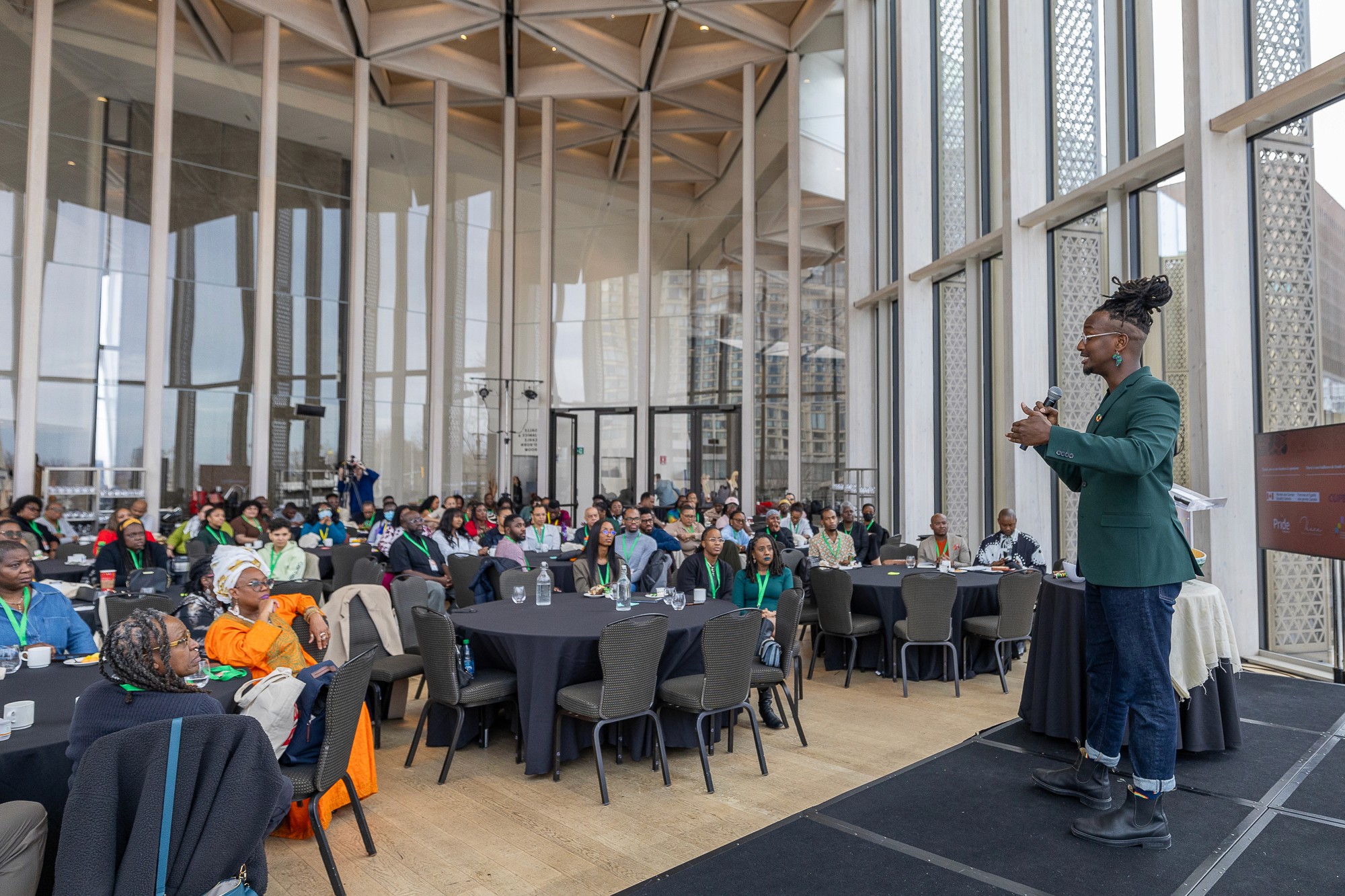 A speaker stands on a stage in a spacious, bright room filled with empty tables and large windows.