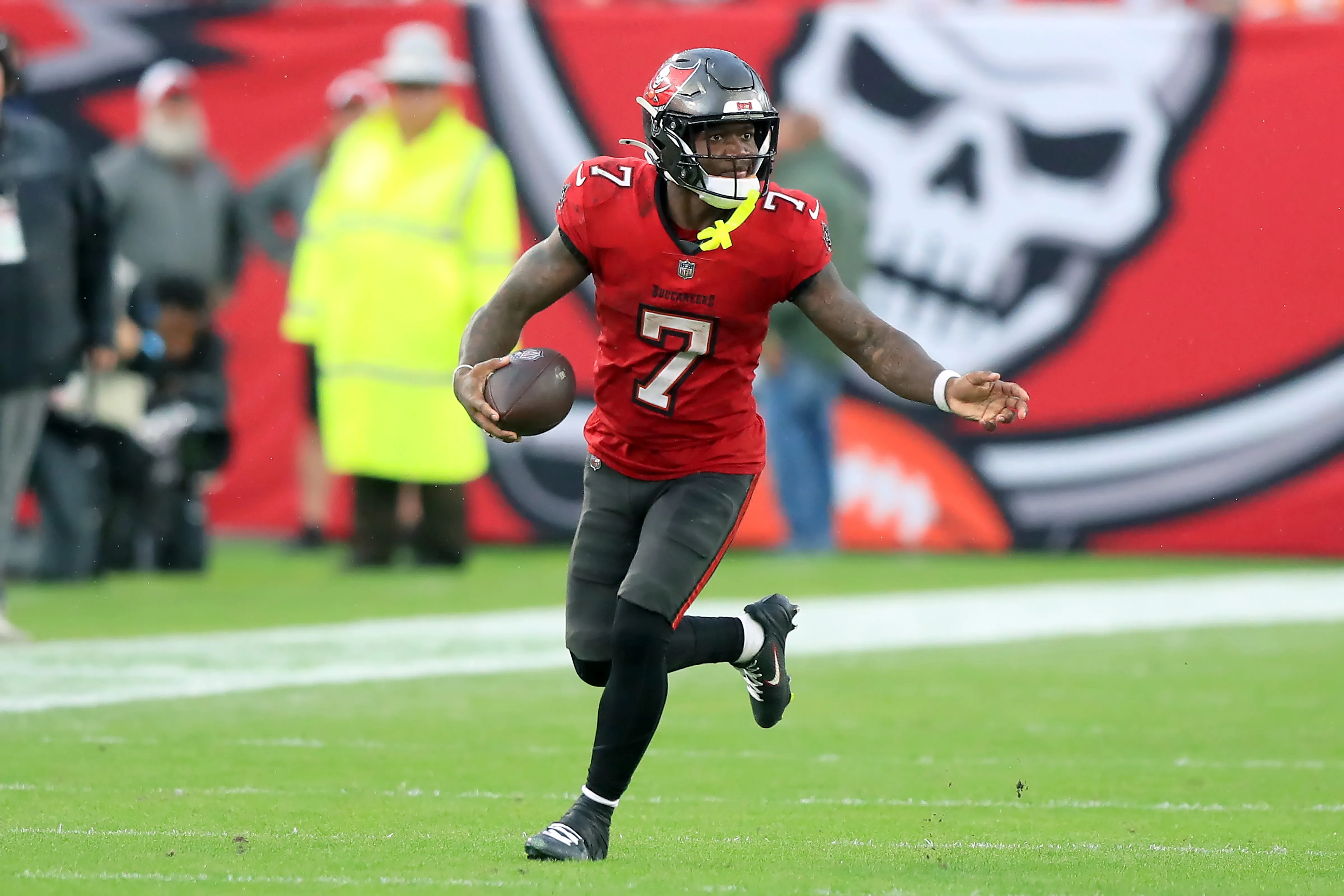 Tampa Bay Buccaneers quarterback wearing number 7 runs with the football on the field, with the team’s pirate flag logo visible on the stadium wall behind him.