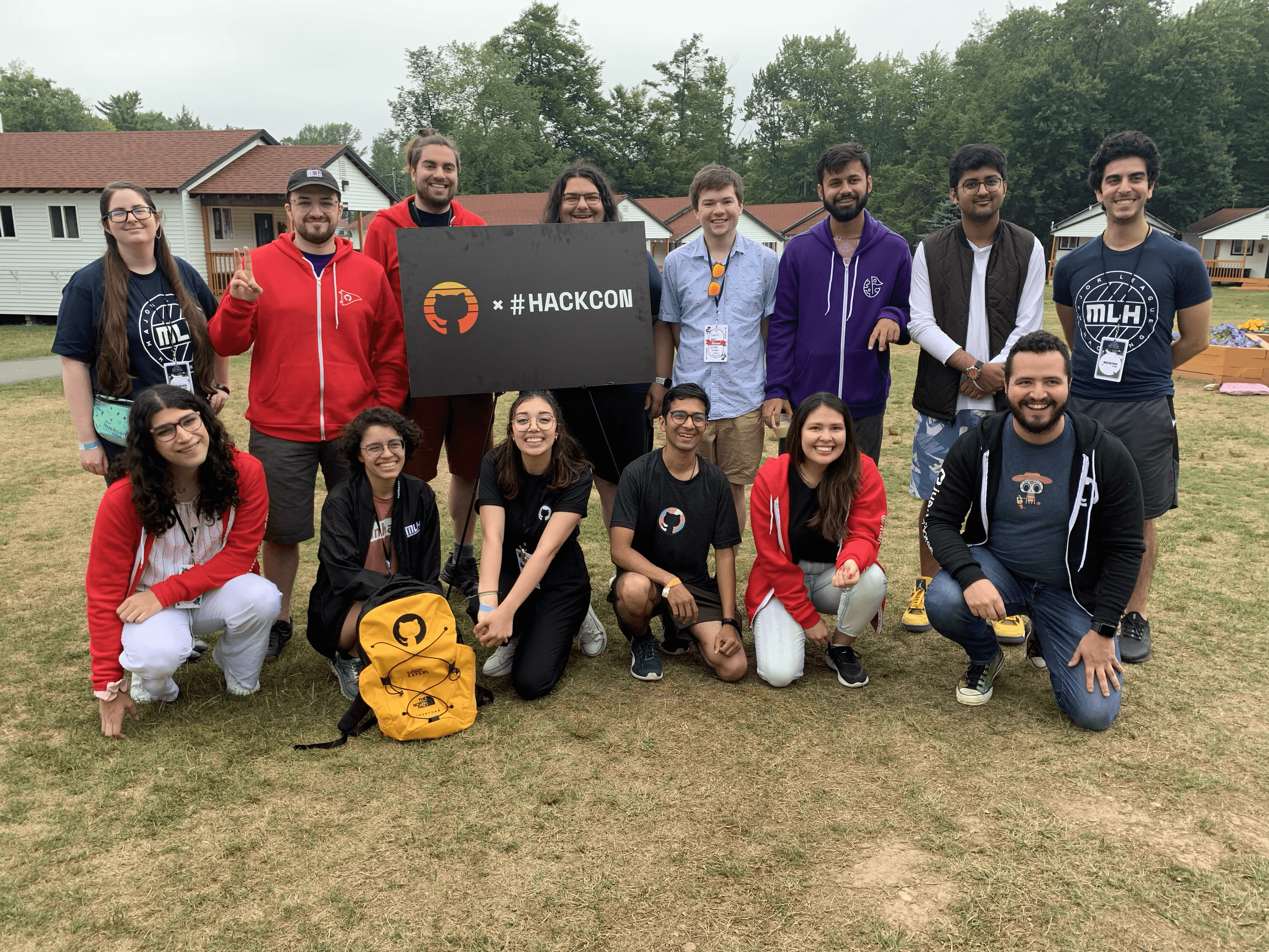 A group people standing in grassy field around a sign with GitHub's octocat logo "x #hackcon".