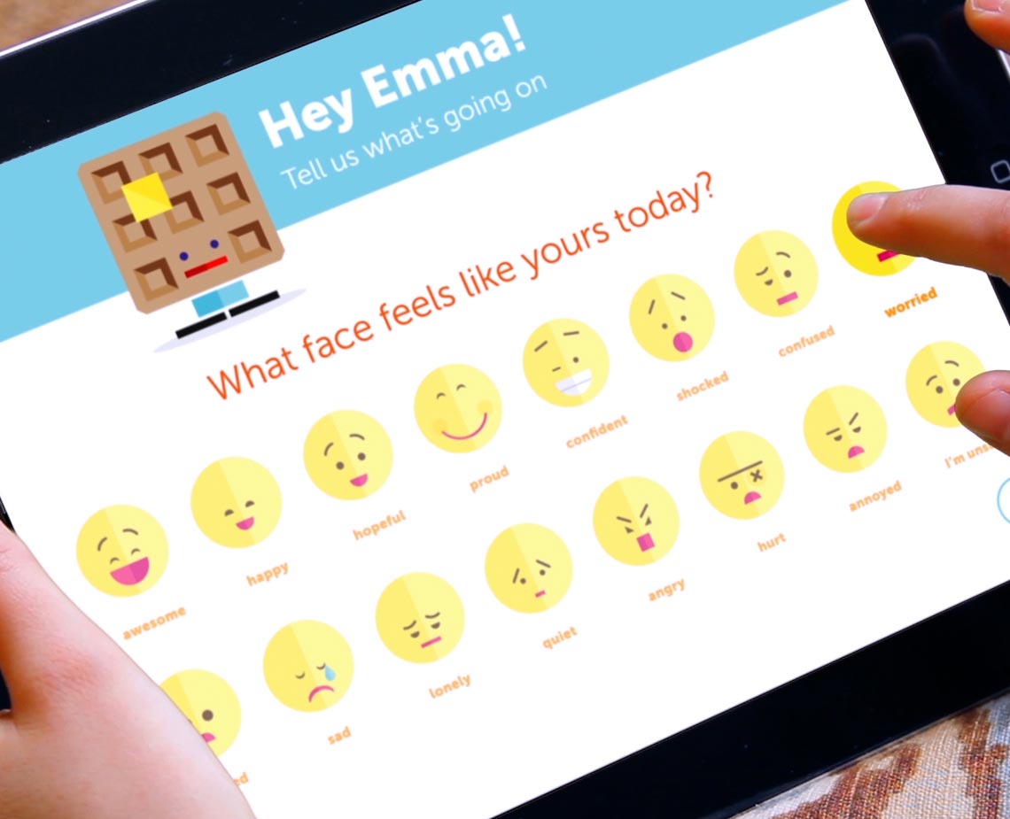 A tablet screen displays a colorful cartoon face emoji grid on a white background. Above the grid, the text “Hey Emma! Tell us what’s going on” appears in blue. Below the grid, the question “What face feels like yours today?” is posed. A hand interacts with the screen on the right side.