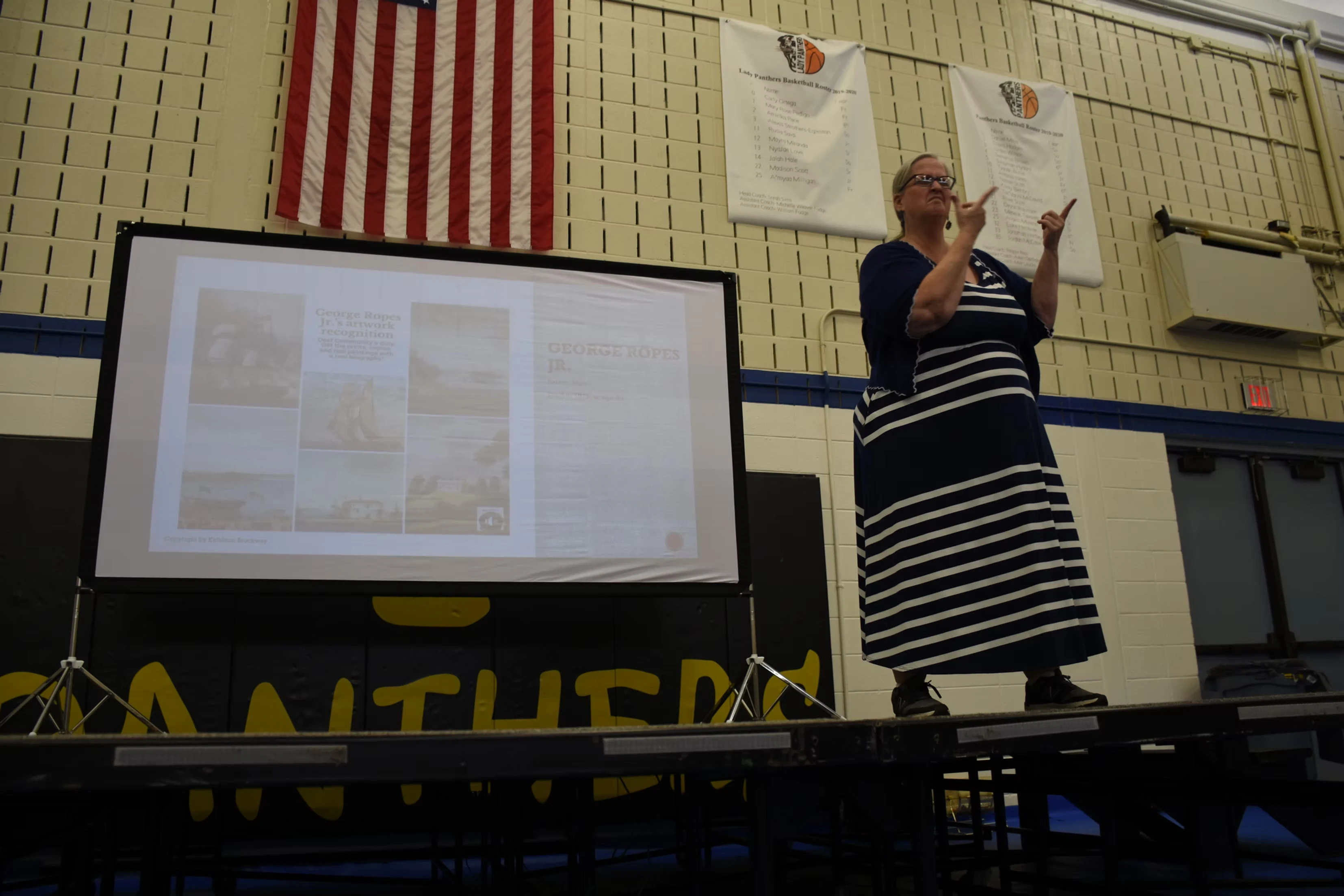 A woman in a striped dress gives a presentation, using sign language, in front of a projection screen showing images.