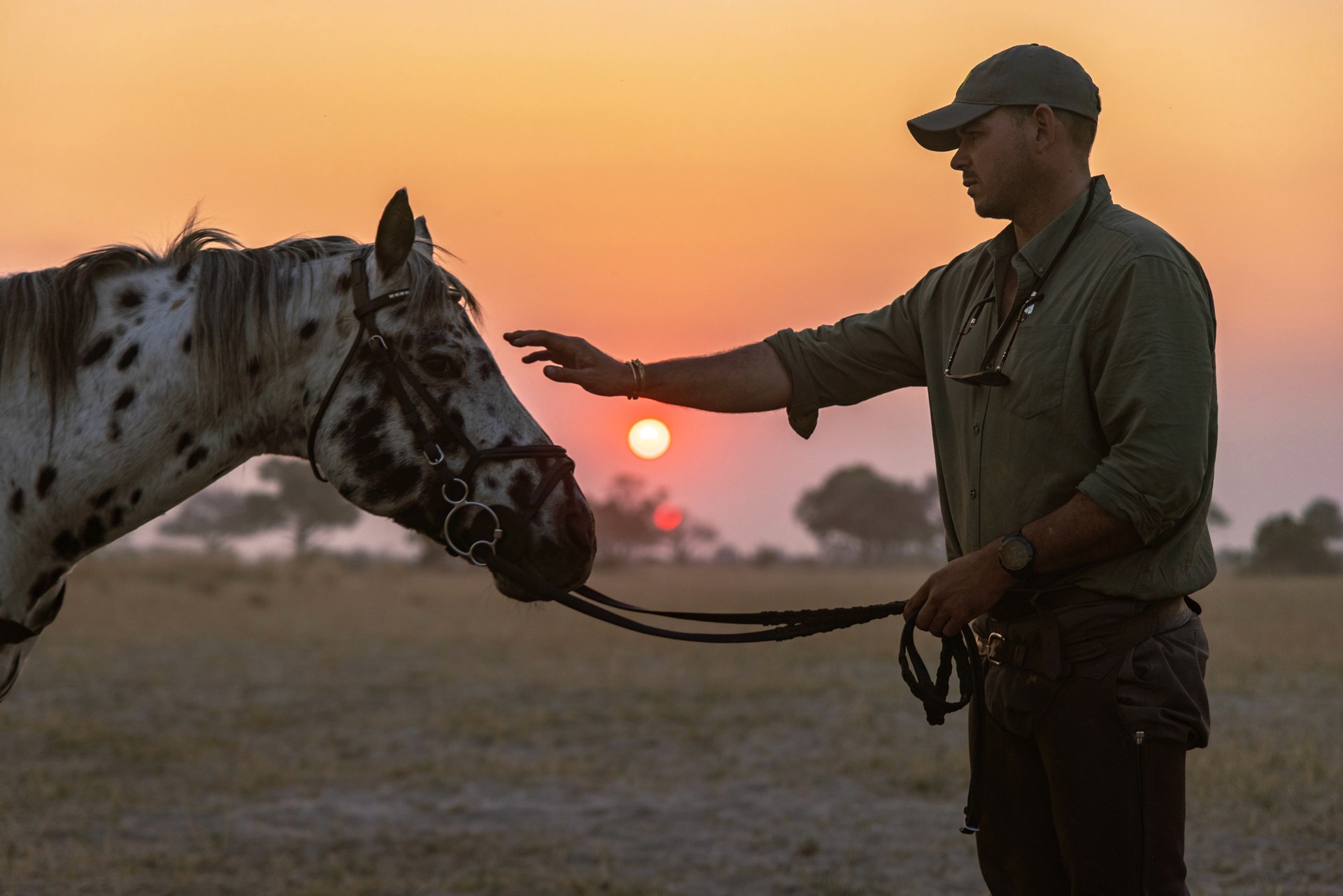 Kujwana Delta Ride, Okavango Delta, Botswana. A horseback safari guide gently stroking the forehead of his horse in the sunset.