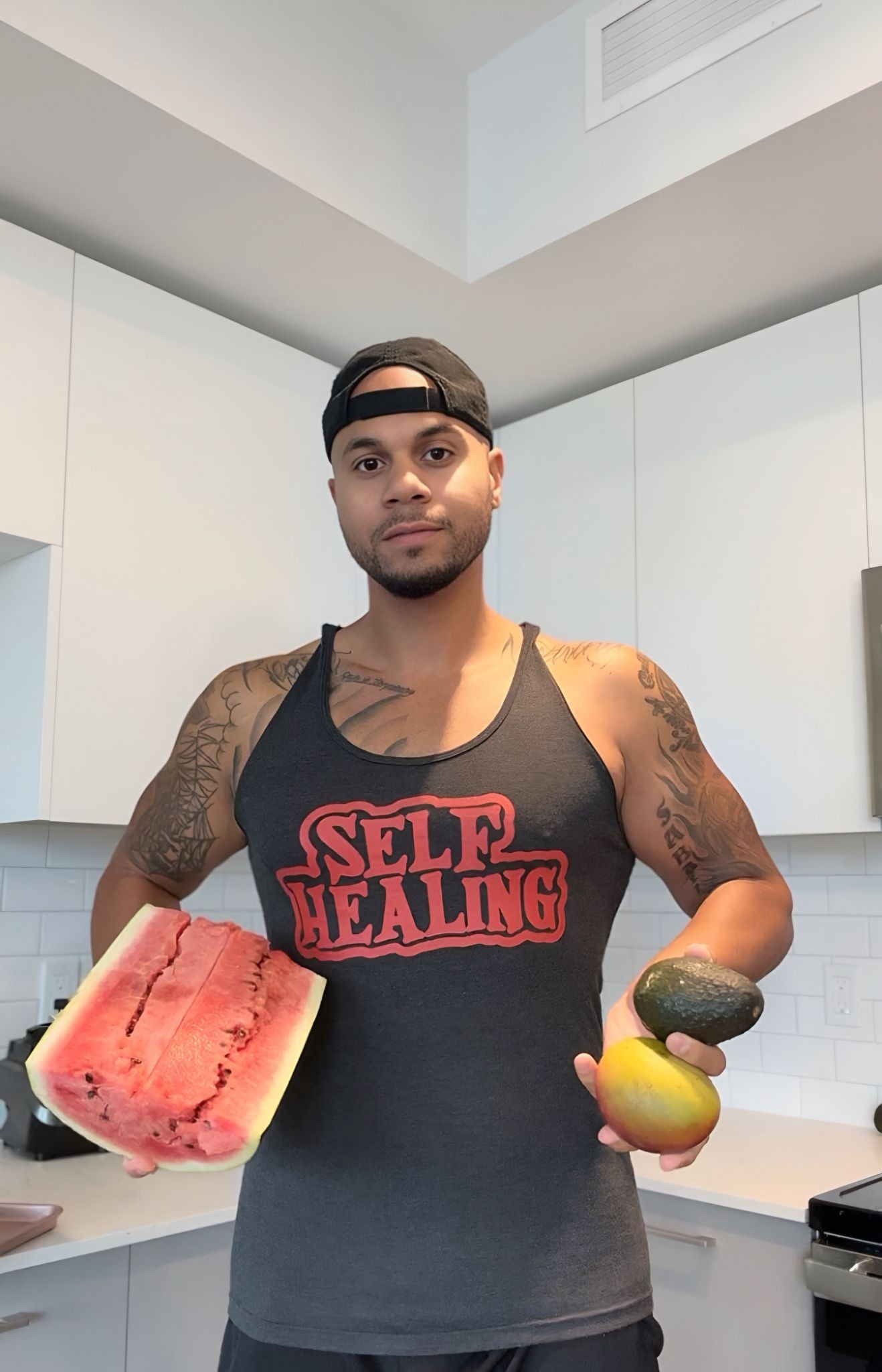 Man in a cap and a tank top with "Self Healing" text, holds watermelon, avocado, and mango in a white kitchen, conveying a health-conscious vibe.