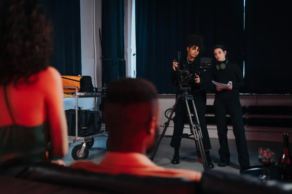 Two crew members operate a camera and check a script in a dark studio while filming two actors from behind.