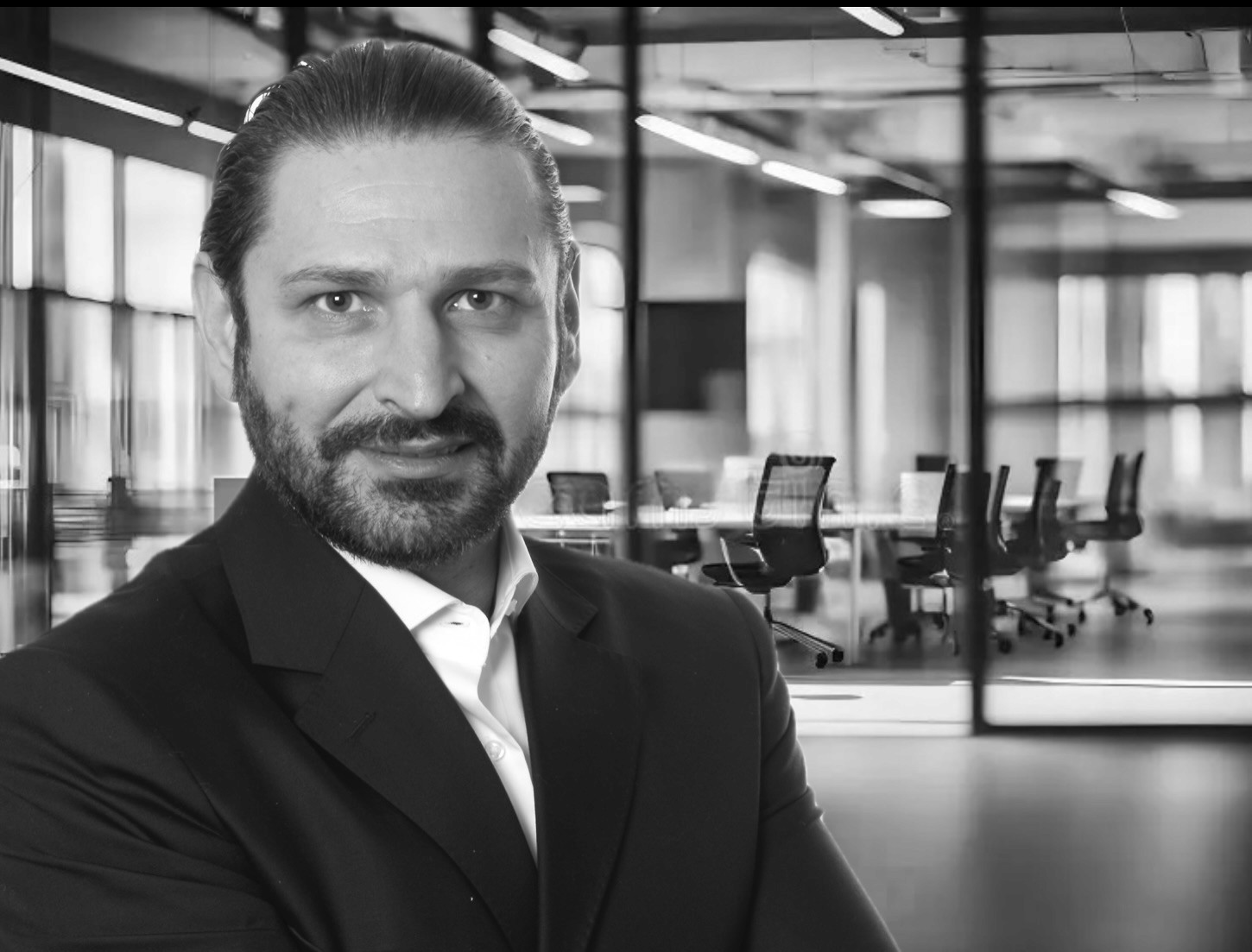 Black and white photo of caucazian man with beard dreassed in a black suit in front of a conference room