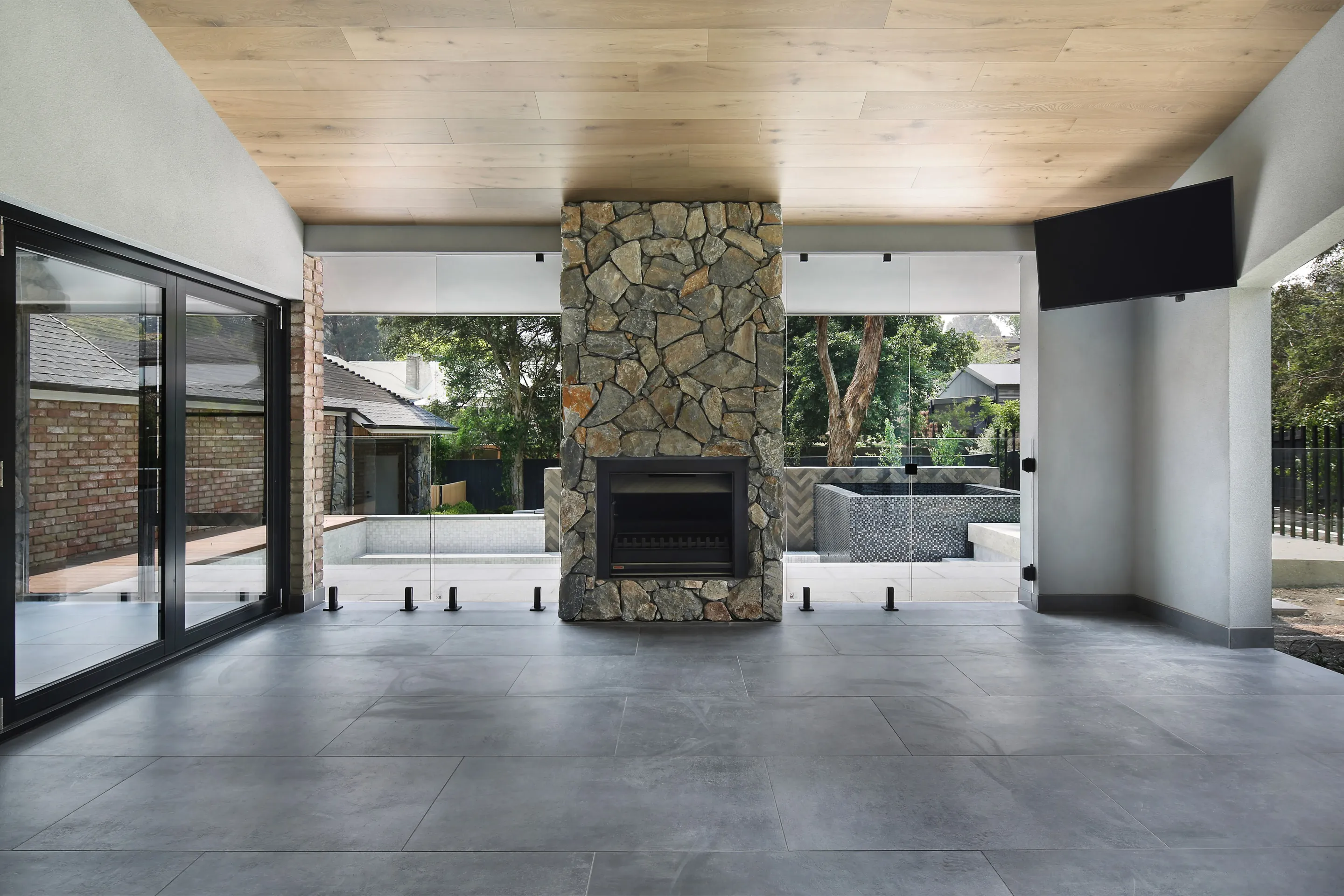 Covered alfresco area with a timber-lined ceiling, a central stacked-stone fireplace flanked by glass framing the pool and spa, and large charcoal tiled floors