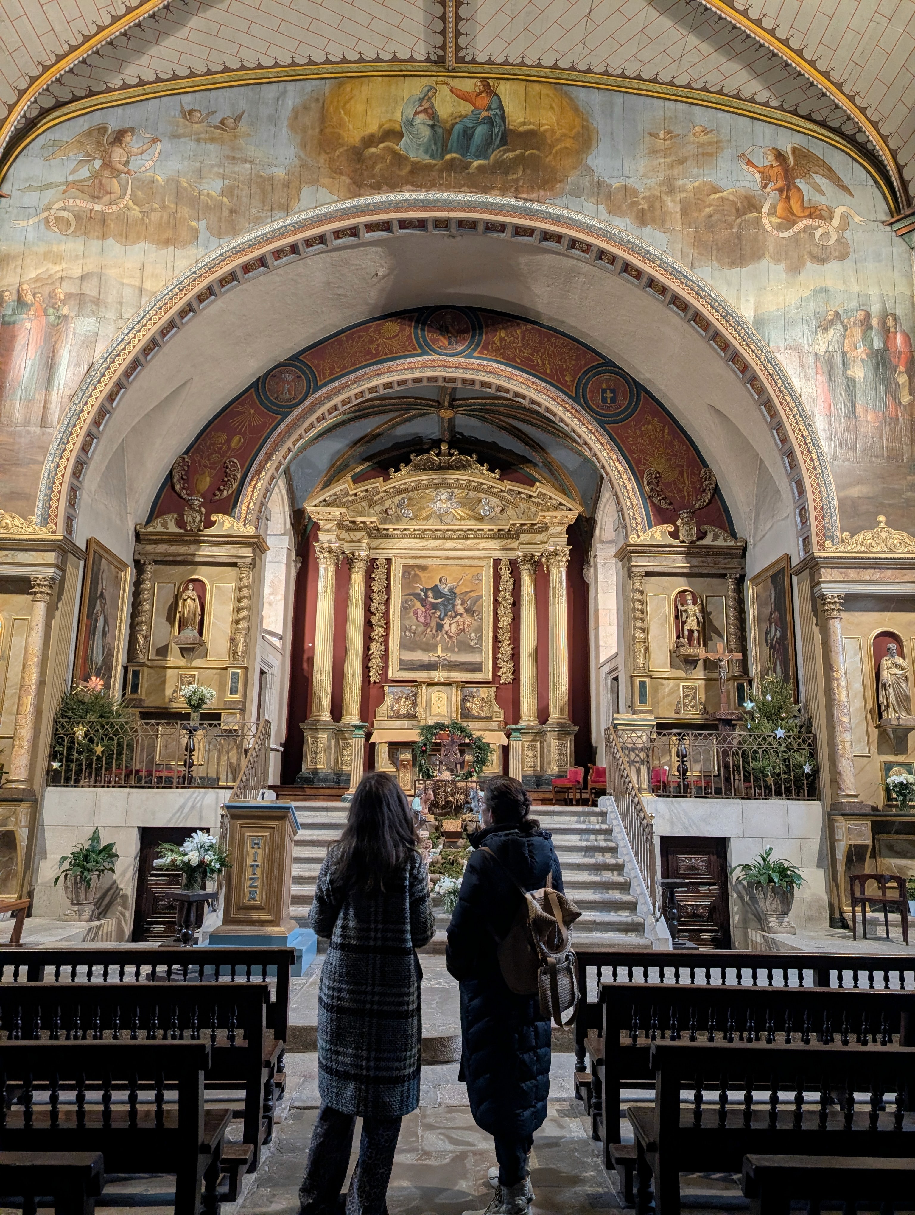 Basque church interior with a gilded altarpiece and frescoes, visitors looking toward the altar, in Sare.
