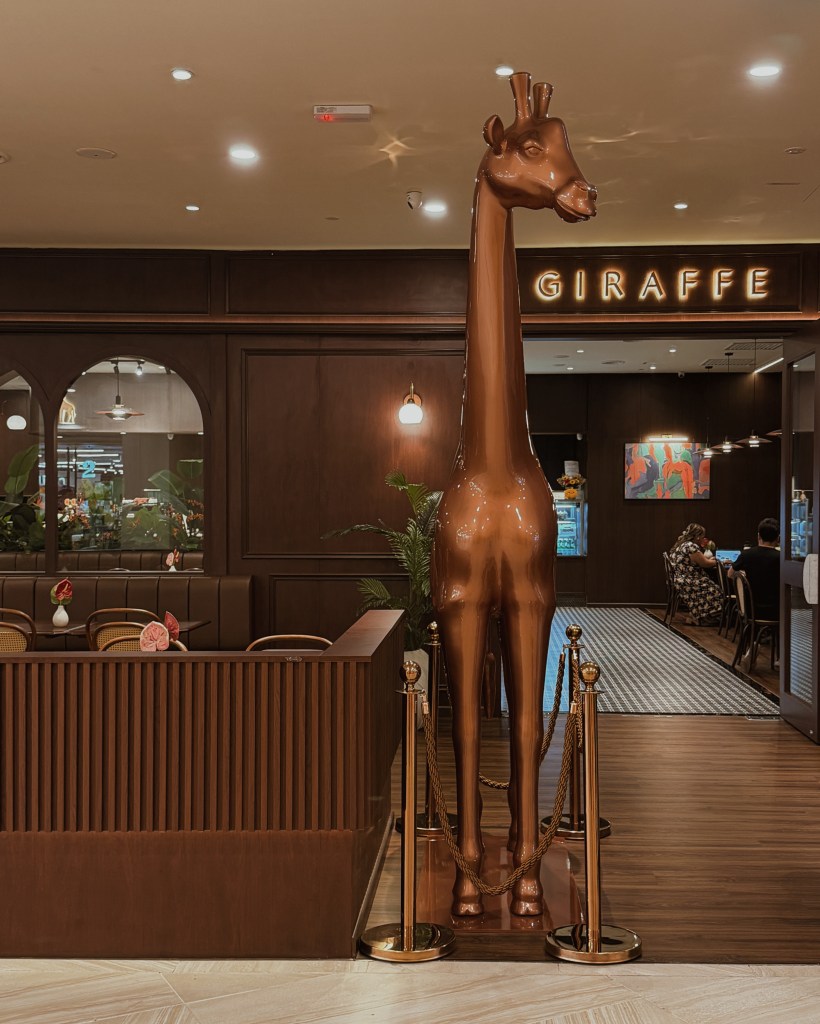 A tall, brown giraffe statue standing near a restaurant entrance with the sign 'GIRAFFE' illuminated in the background.