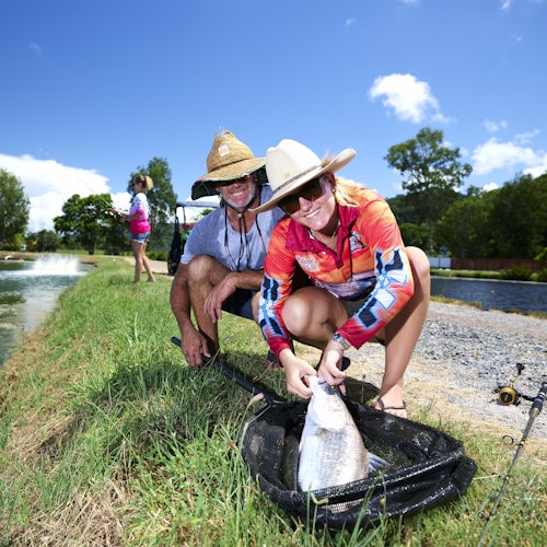 在一个阳光明媚的日子里,两个人戴着草帽在池塘边举着一条大鱼,旁边的地上放着渔具。