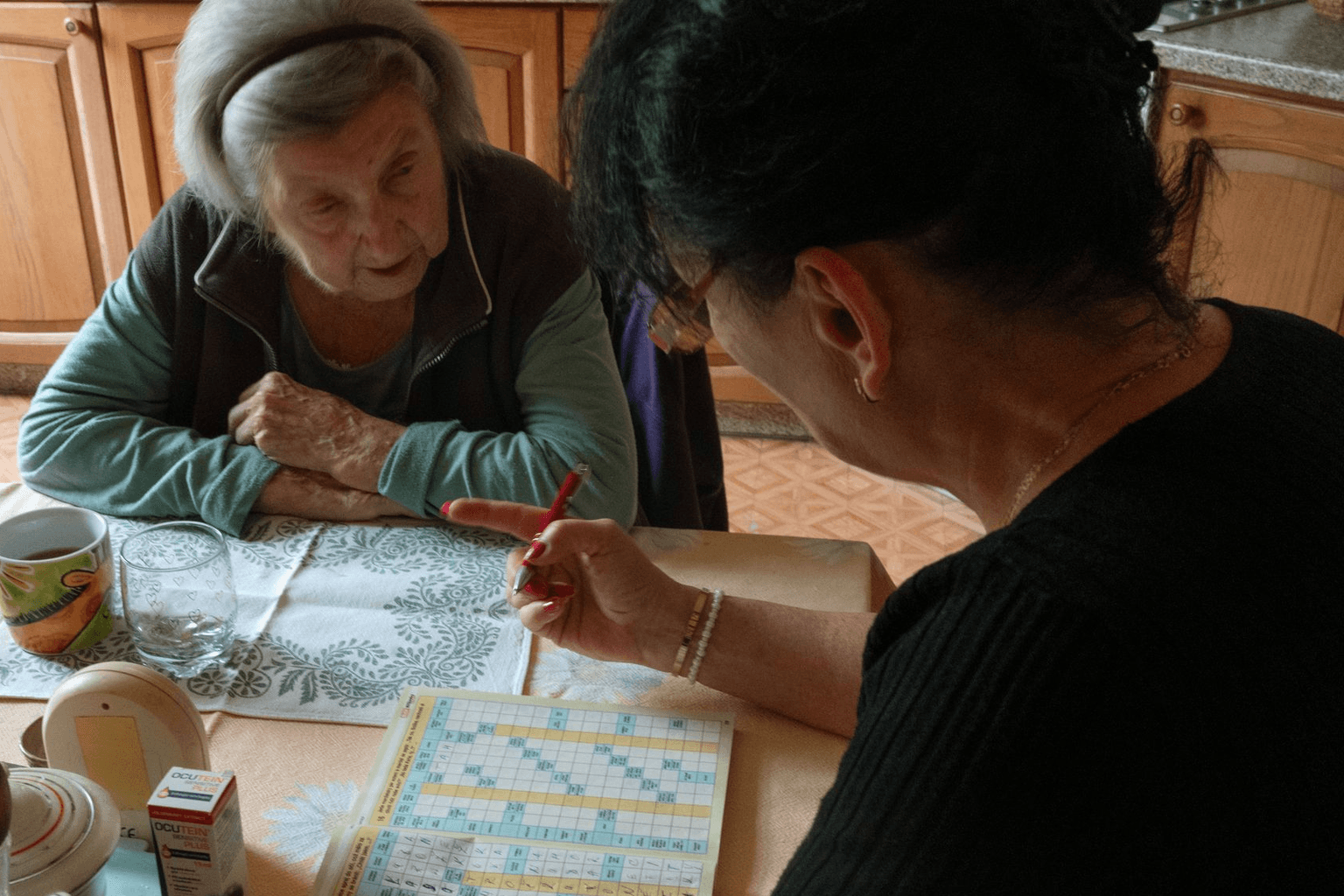 An older woman and a younger woman working through a crossword together at her kitchen table.